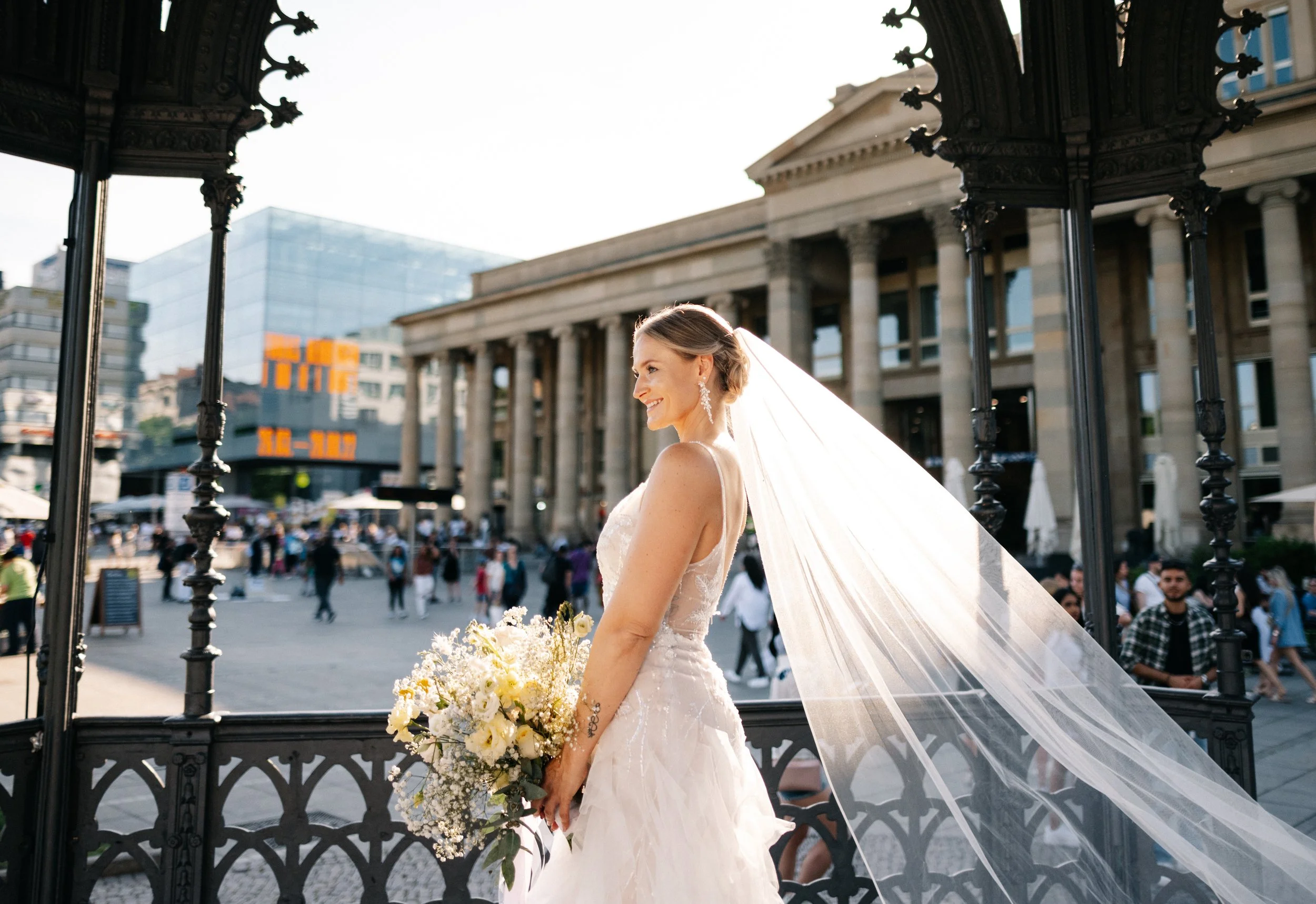 Braut in Hochzeitskleid mit Blumenstrauß auf einer Stadtbrüstung, im Hintergrund Menschen und Gebäude, Sonnenlicht, fröhliche Atmosphäre.