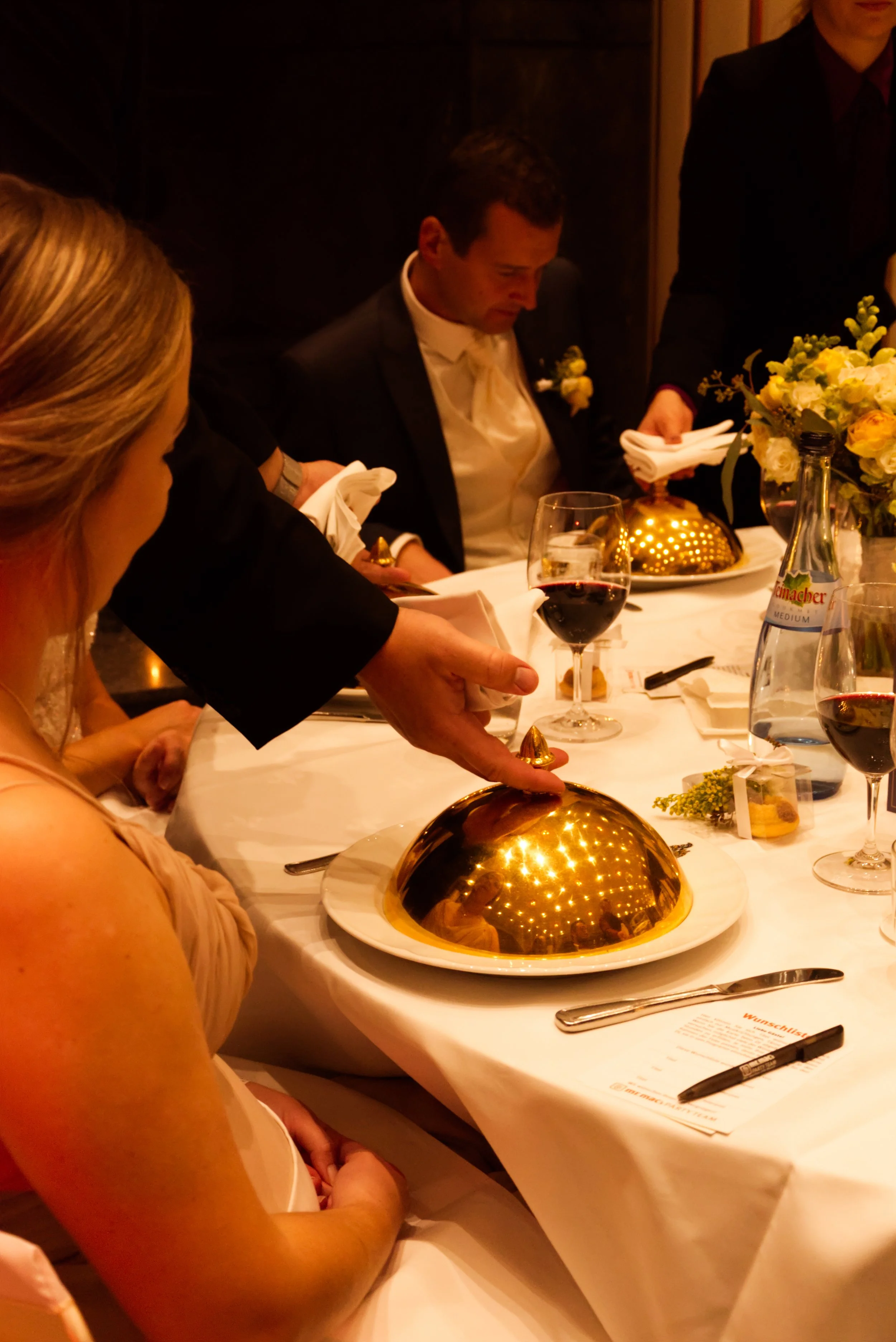 Jemand hebt einen goldenen Deckel von einem Gericht bei einer formellen Dinnerveranstaltung in einem eleganten Raum abgedeckt.
