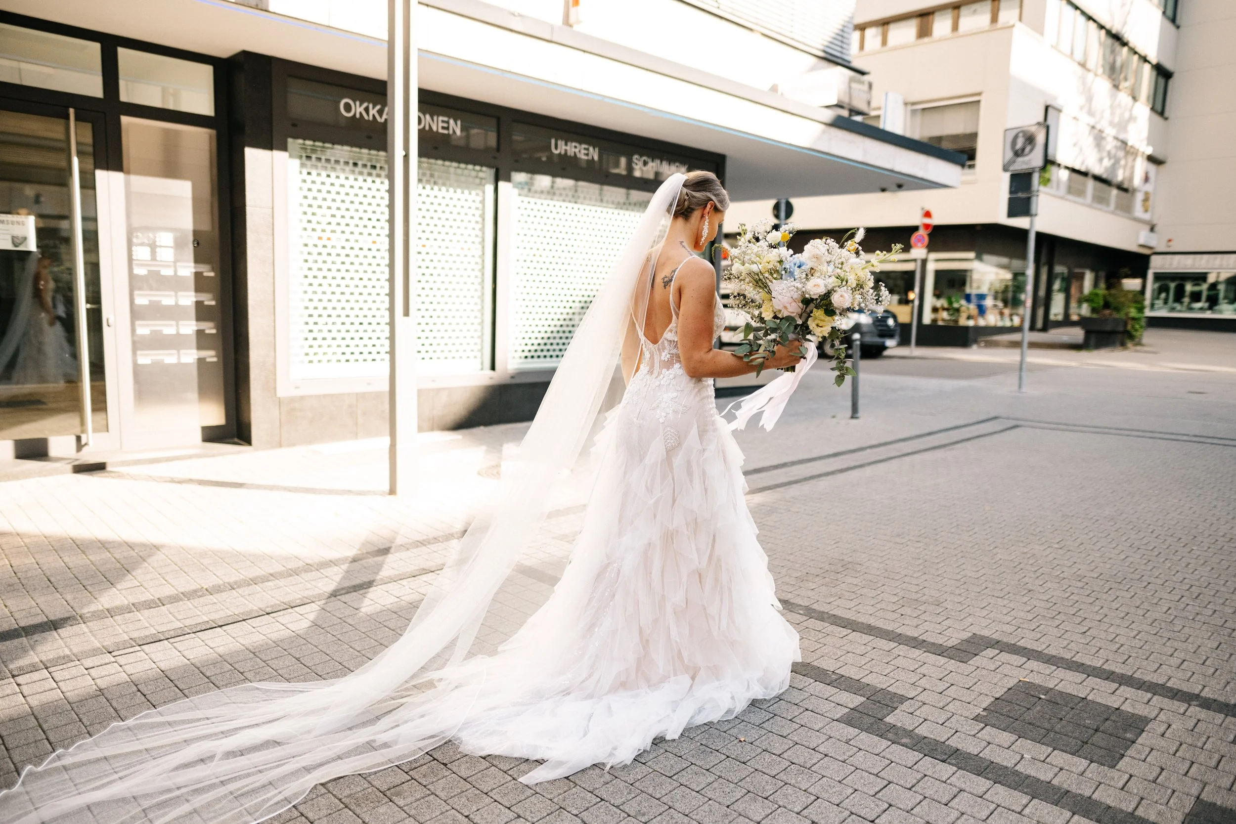 Braut in Hochzeitskleid mit langem Schleier und Blumenstrauß auf Stadtstraßen