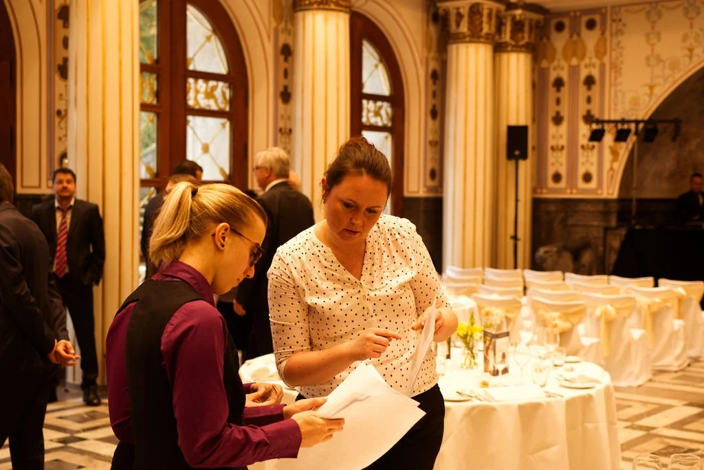 Sieben Menschen in einem eleganten, historischen Saal mit hohen Fenstern, die sich mit weißen und goldenen Dekorationen schmücken. Zwei Frauen im Vordergrund sprechen miteinander und halten Papiere. Weitere Personen im Hintergrund, einige stehen und 