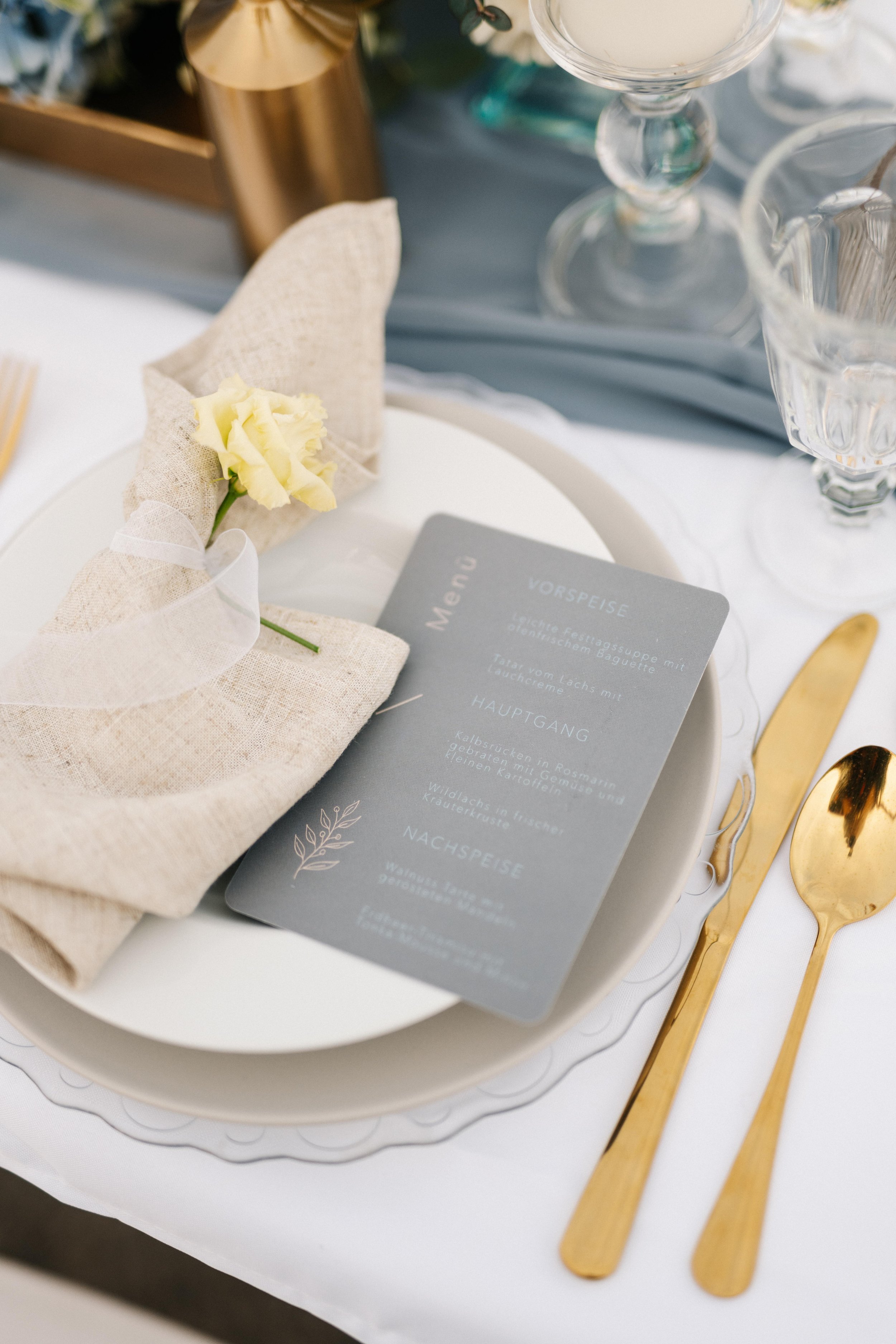 Tischgedeck mit einer weißen Schale, einer Stoffserviette mit einer gelben Blume, goldene Messerklinge und Löffel, einem Menükarte, mehreren Gläsern, und einer grauen Tischdecke im Hintergrund.