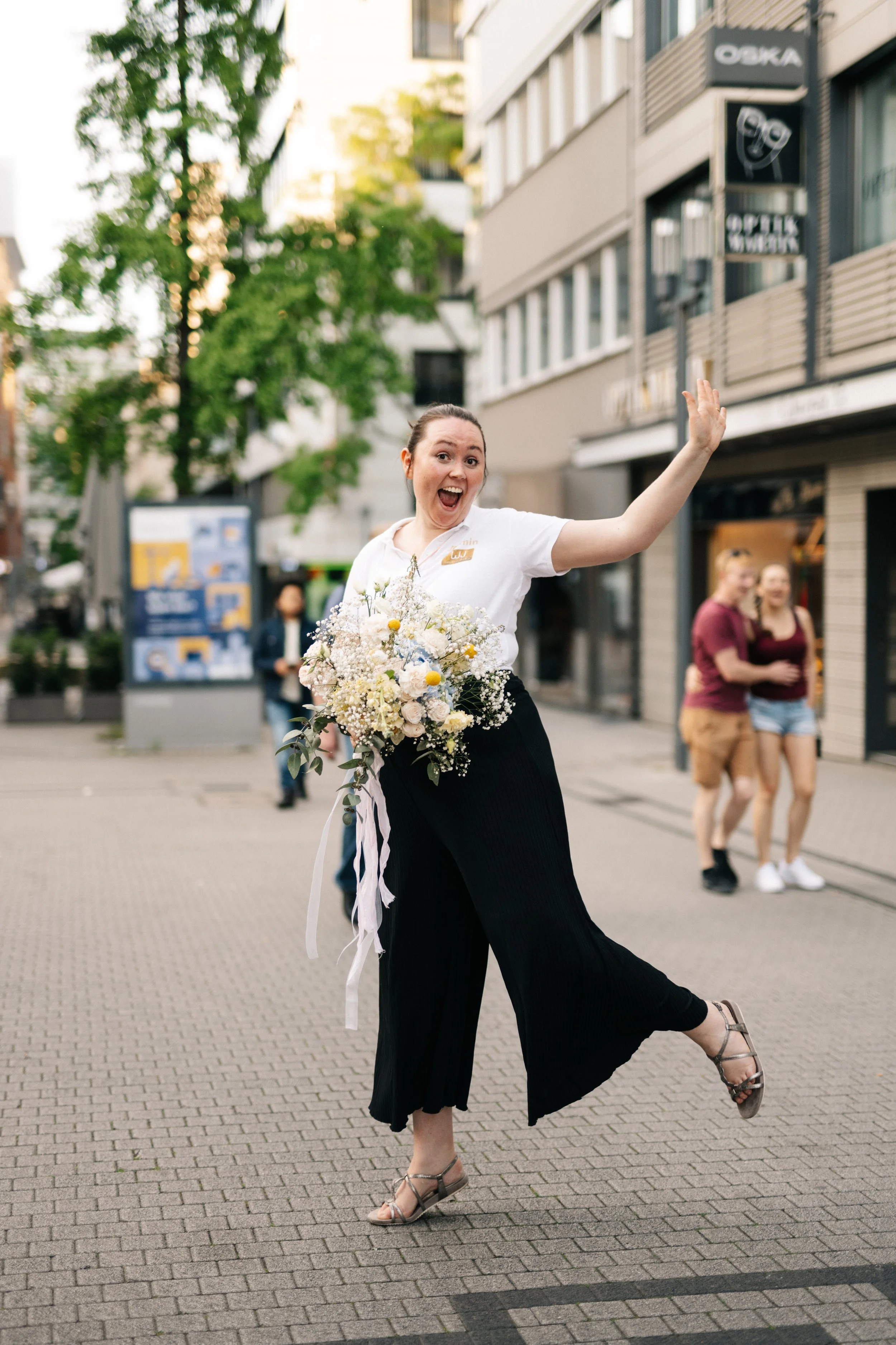 Freudige Frau hält Blumenstrauß in der Hand, posiert auf Straße, lächelt und winkt, im Hintergrund moderne Gebäude und zwei lachende Frauen.