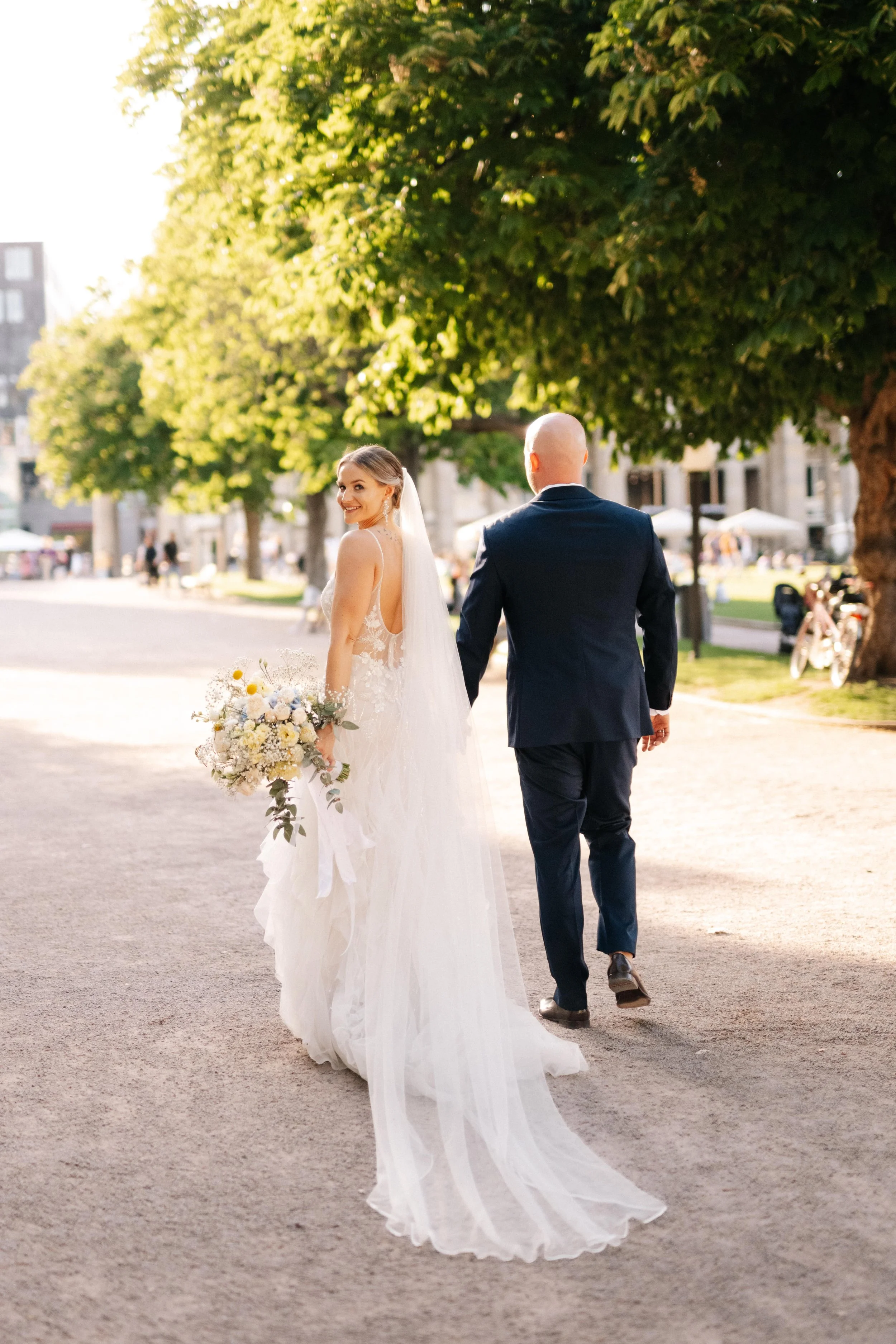 Braut in weißem Kleid und langem Schleier mit Brautstrauß, Rammler in dunklem Anzug, Spaziergang im Park an sonnigem Nachmittag, grüne Bäume im Hintergrund.