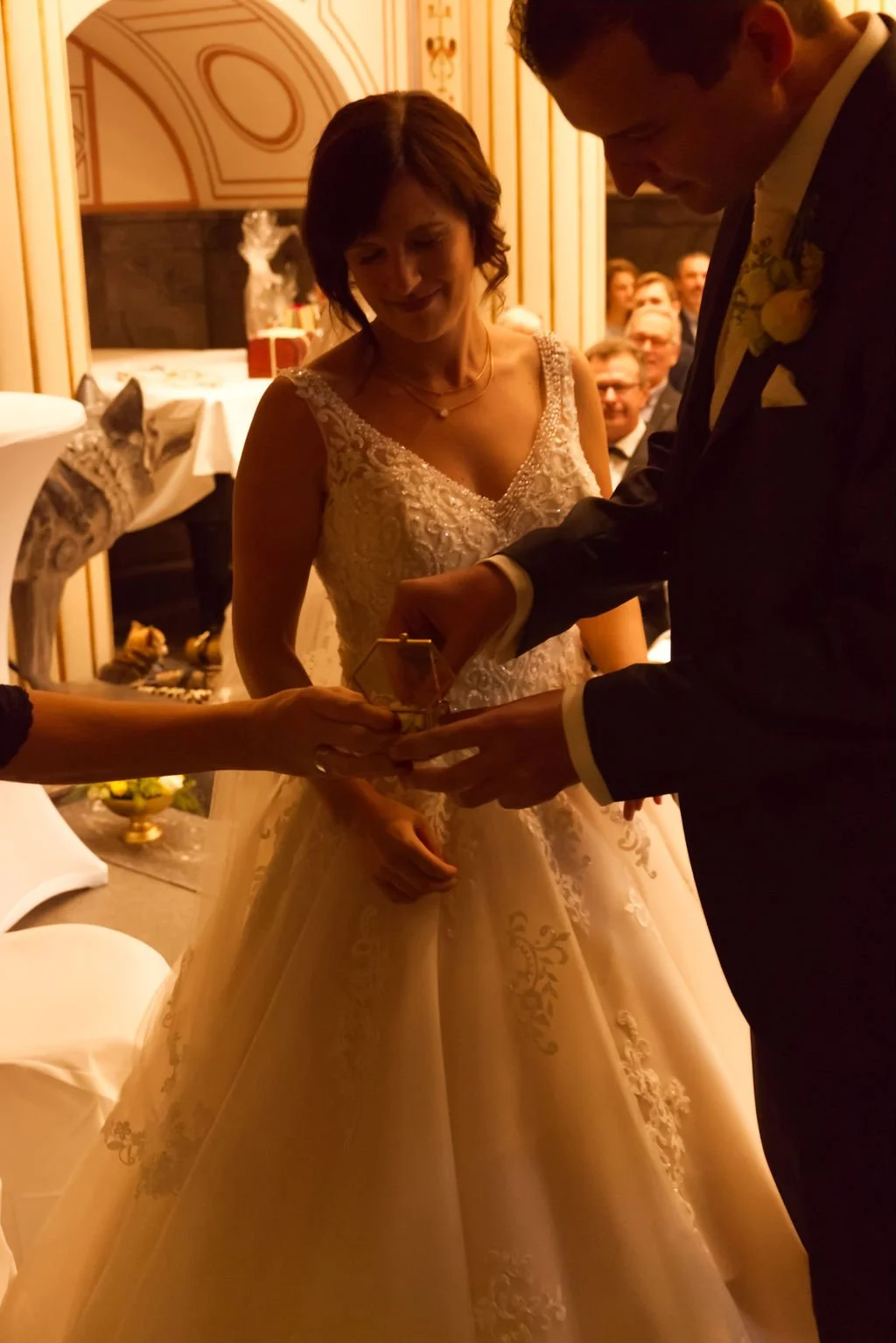 Braut und Bräutigam bei der Hochzeitszeremonie, während sie einen Ring austauschen, in einem festlich dekorierten Raum mit Gästen im Hintergrund.