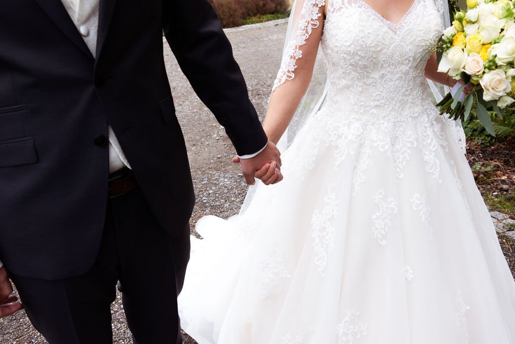 Braut und Bräutigam halten sich an den Händen, Braut trägt ein weißes, schlichtes Hochzeitskleid mit Spitze und hält einen Blumenstrauß.