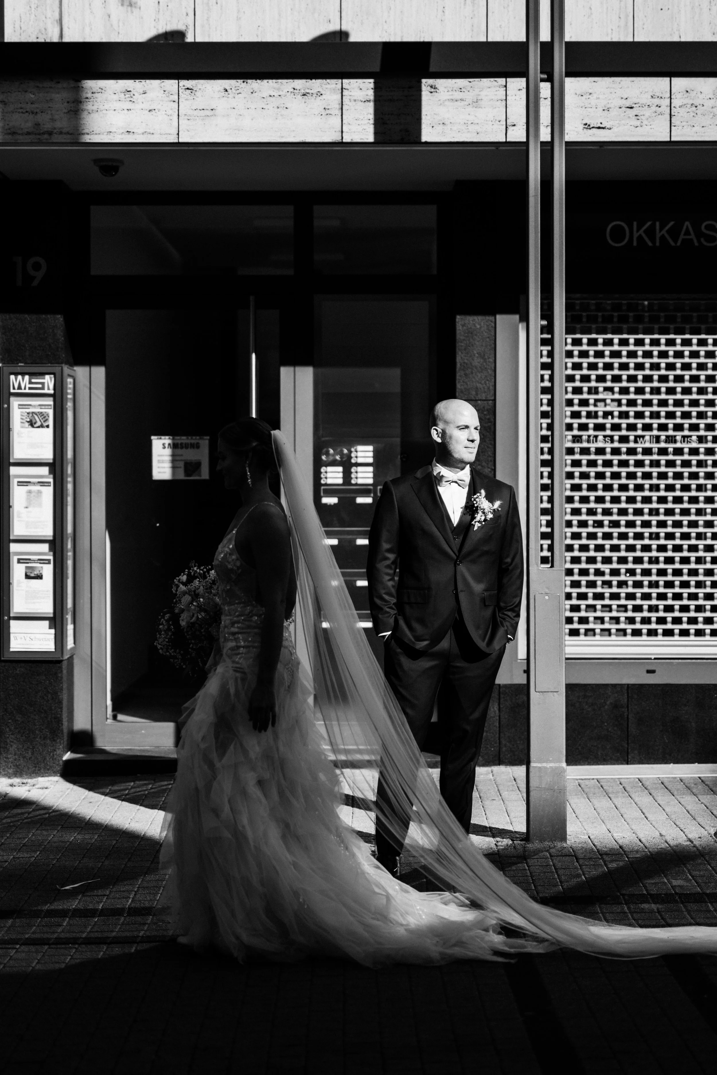 Paare in Hochzeitskleidung stehen vor einem Gebäude, dabei eine Frau in Brautkleid mit Schleier und ein Mann in Anzug, Schwarz-Weiß-Foto.