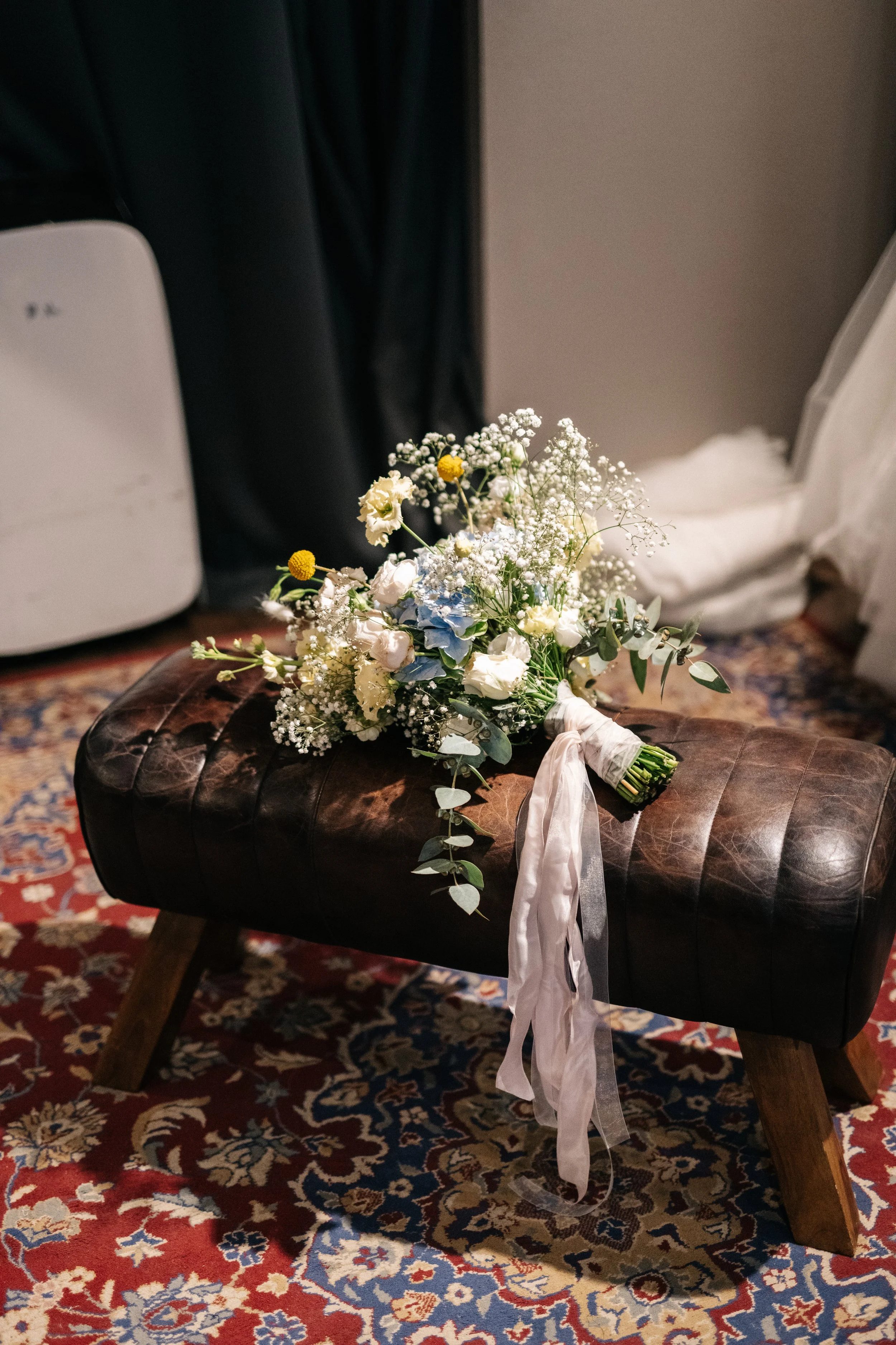 Brautstrauß mit weißen, blauen und kleinen gelben Blumen, liegt auf einem braunen, gepolsterten Hocker auf einem bunten Teppich.