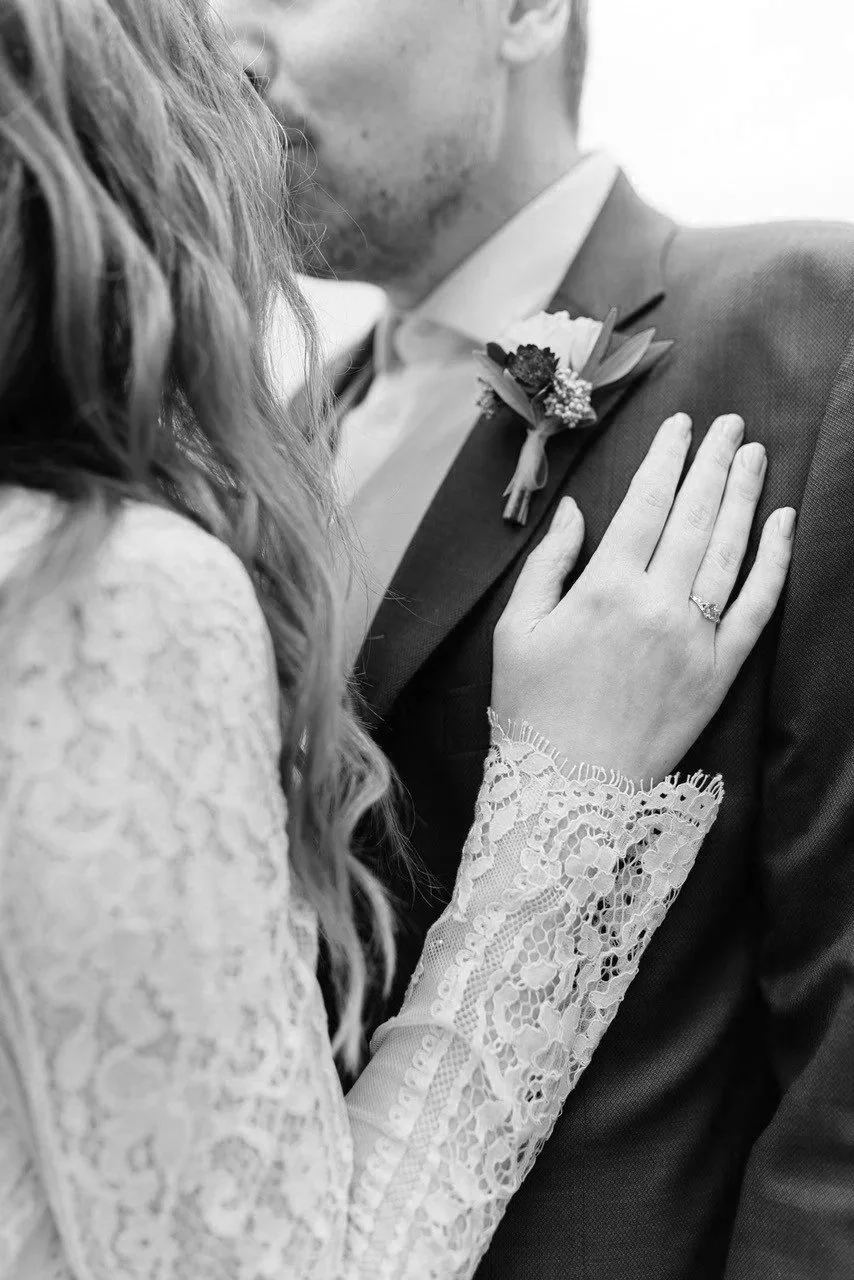 Nahaufnahme einer Braut, die ihre Hand mit Ehering auf die Brust eines Mannes legt. Sie trägt ein Spitzenkleid und eine Blume im Haar. Das Bild ist schwarz-weiß.