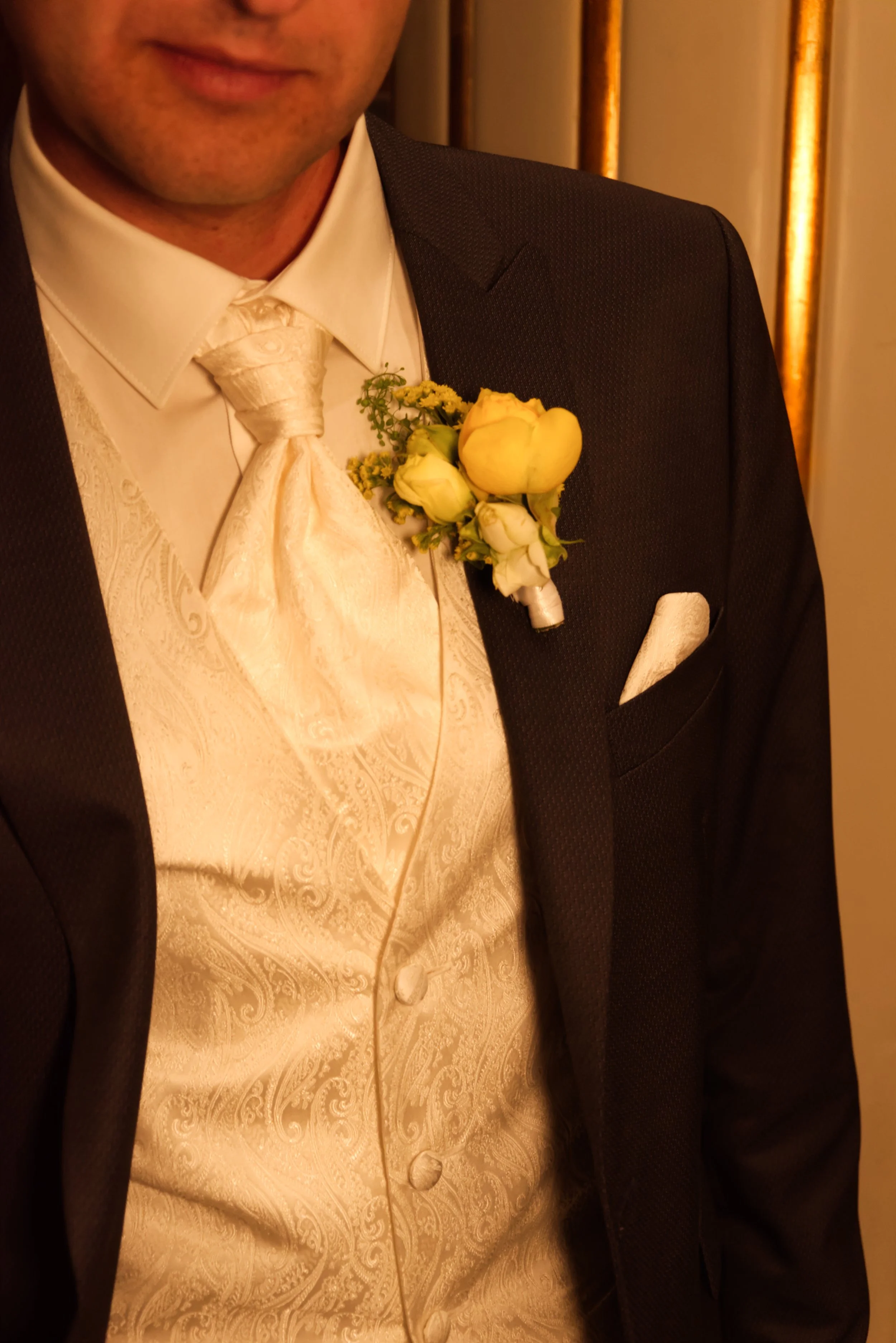 Ein Mann trägt einen schwarzen Anzug mit weißer Fliege, einer weißen Weste mit Maserung und hat eine Orchideen-Brosche am Revers, passend zur Hochzeit.