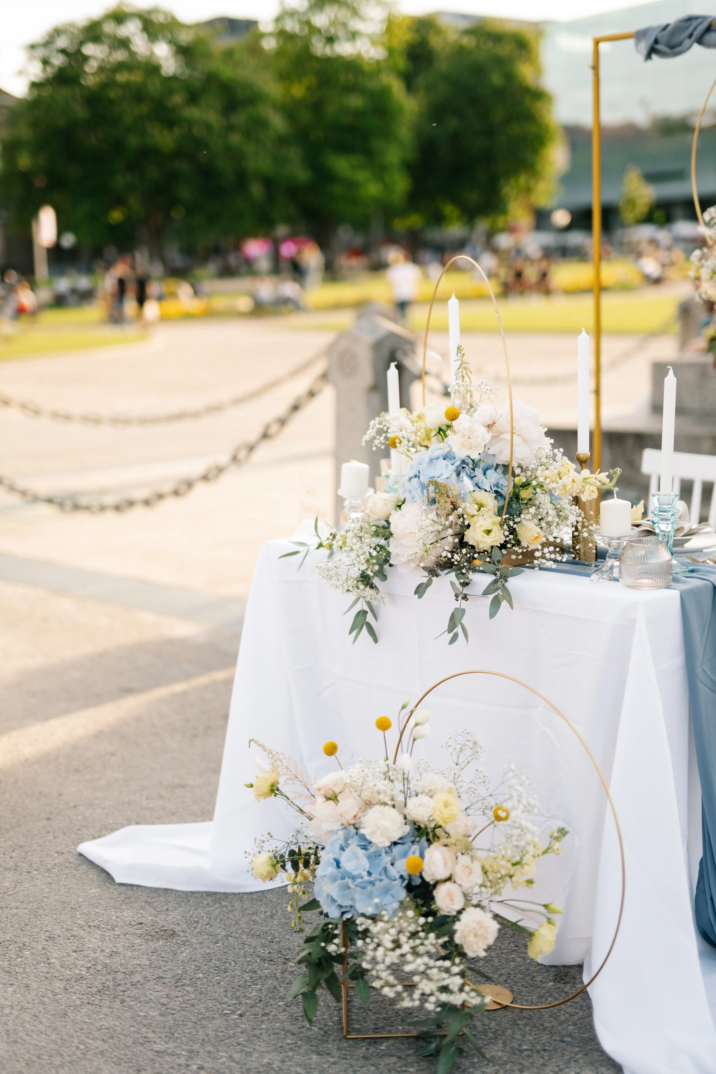 Blumendekoration mit weißen, blauen und gelben Blumen auf einem weißen Tischtuch bei einer Outdoor-Veranstaltung, vermutlich Hochzeit im Frühling oder Sommer.