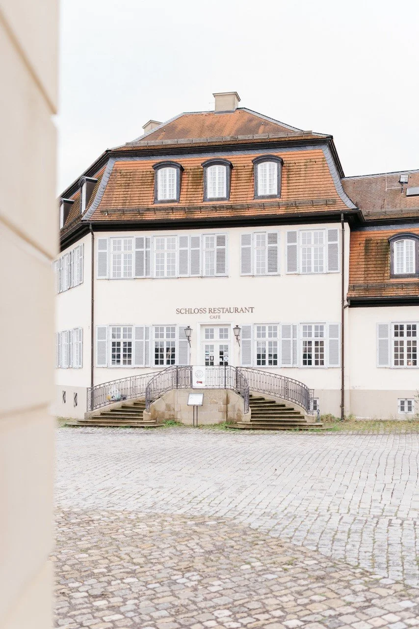 Ein historisches Gebäude mit roten Ziegeldach und weißen Fensterläden, das als Schlossrestaurant und Café bezeichnet wird, mit Treppen vor dem Eingang auf einem gepflasterten Platz.