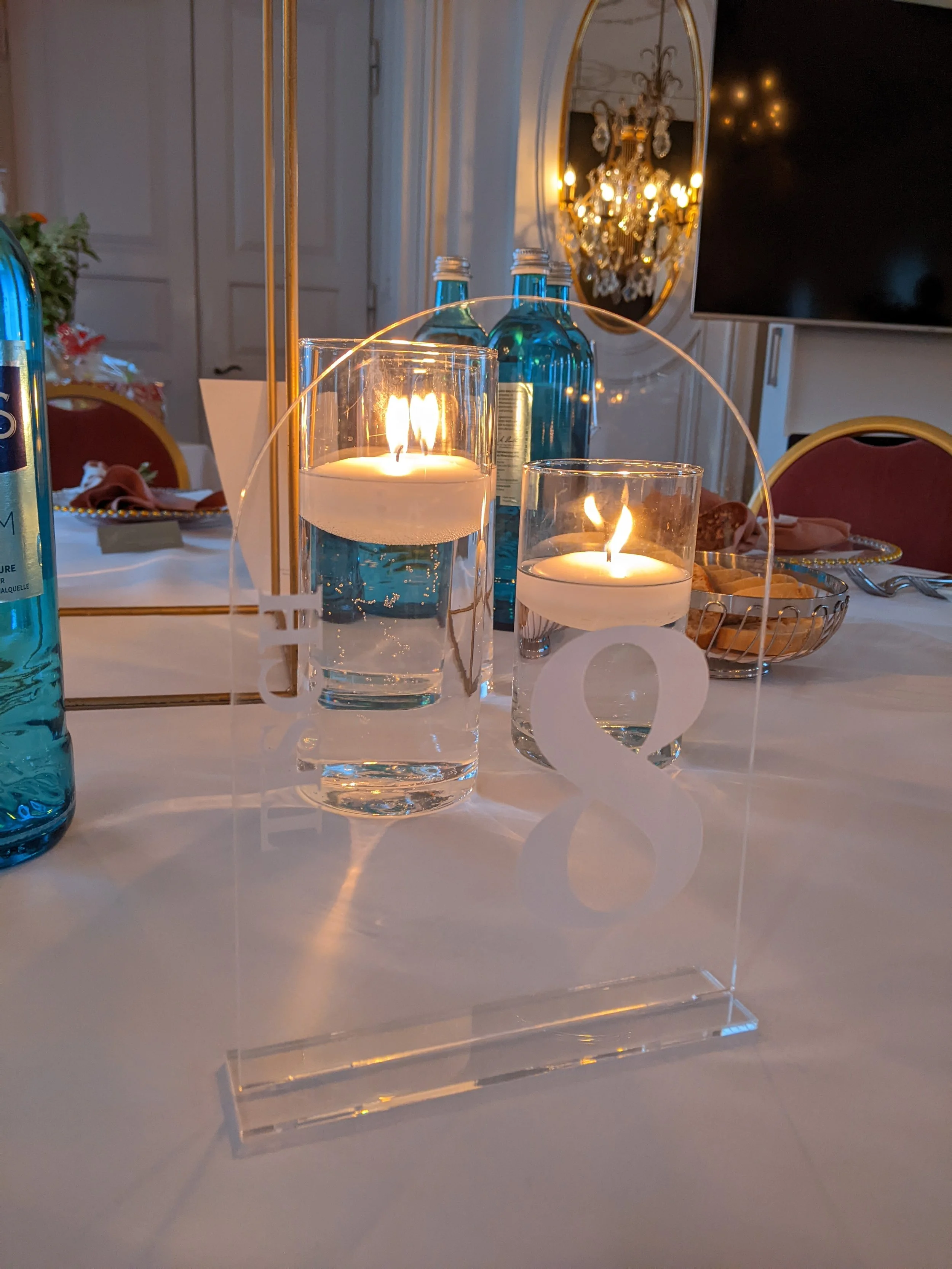 Tischdekoration mit zwei brennenden Kerzen in Wasser gefassten Gläsern, blauen Wasserflaschen, einer Glasscheibe mit Zahl acht, in einem elegant dekorierten Raum mit Kristallleuchter und Fernseher.