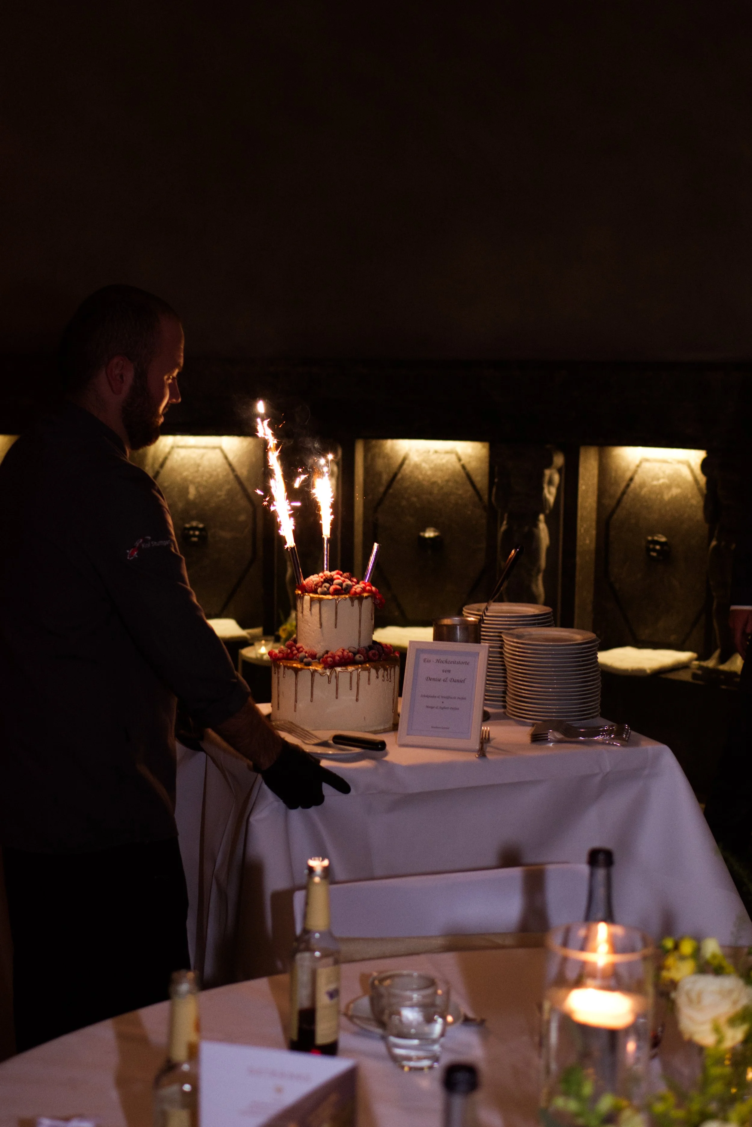 Ein Mann steht bei einer zweistöckigen Hochzeitstorte mit Schokoladenganache und Beeren, umgeben von Kerzen, auf einem weißen Tisch, während Feuerwerkskörper auf der Torte entspringen.