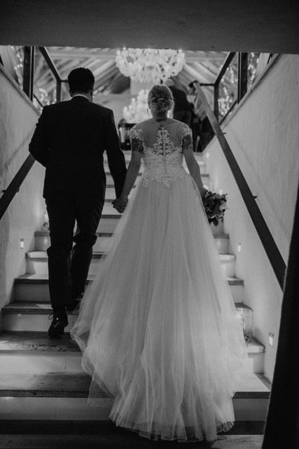 Braut und Bräutigam gehen die Treppe in einem Hochzeitsort hinauf, mit romantischer Beleuchtung und Deckenleuchte im Hintergrund.