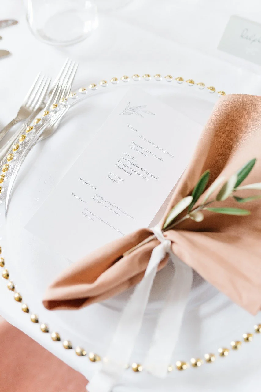 Eleganter weißer Tisch mit goldverzierten Tellern, einem Menü auf weißem Papier, einer rosa Serviette mit grünen Zweigen, Besteck und einem Glas.