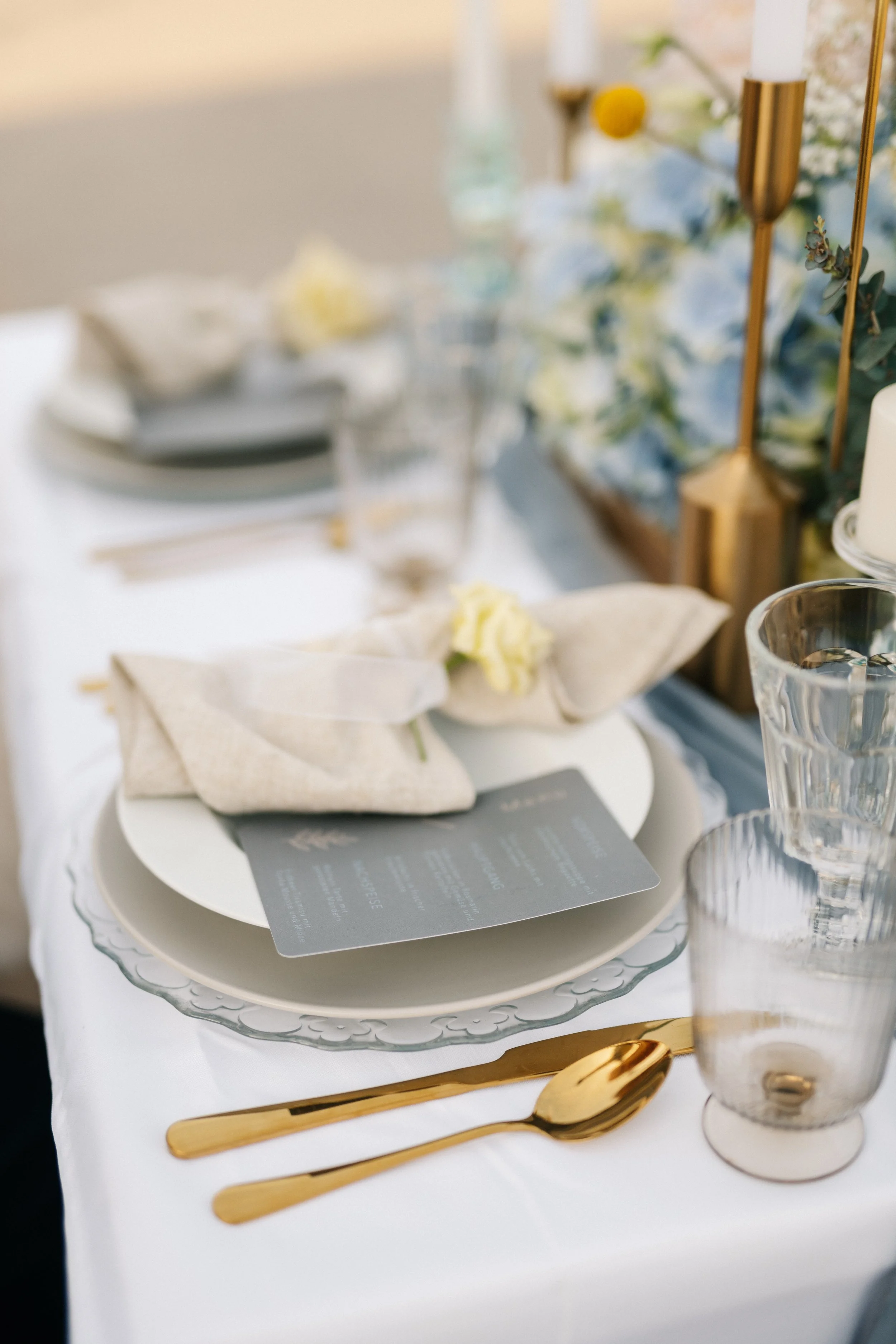 Eleganter gedeckter Tisch mit weißen Tellern, goldenen Besteck, Gläsern und einer floral Dekoration mit blauen und weißen Blumen.
