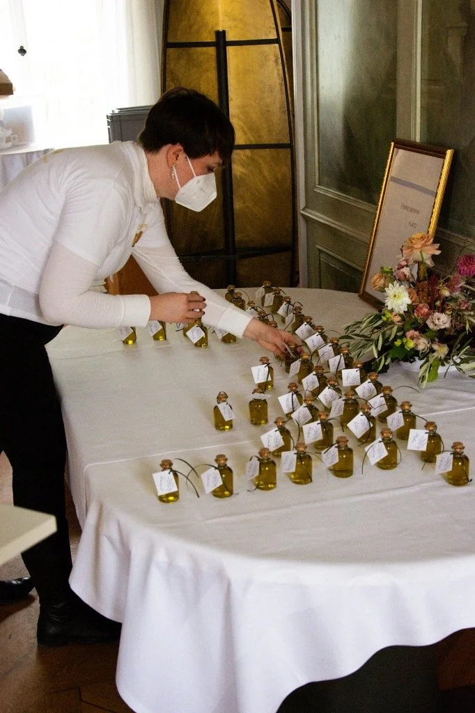 Person in weißen Hemd und Gesichtsmasker richtet kleine Flakons auf einem Tisch mit weißen Tischtuch zu.