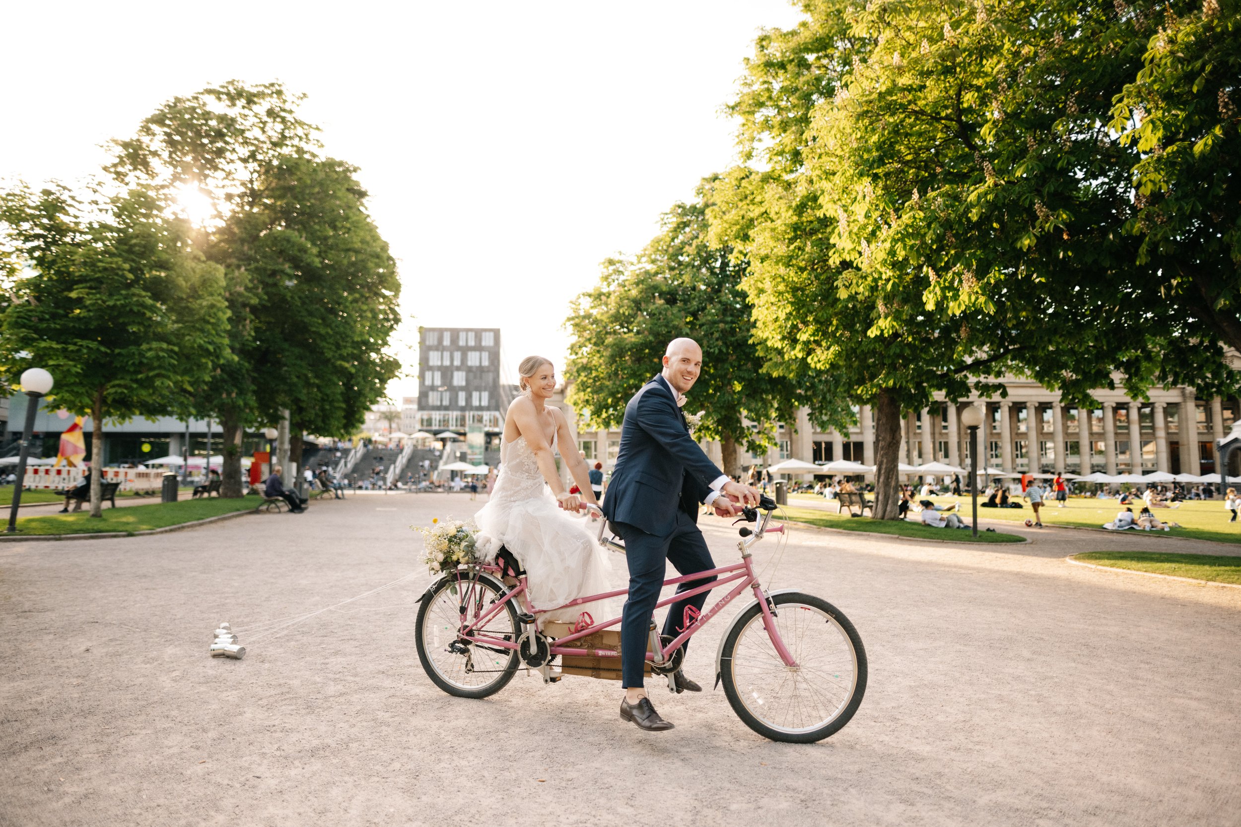 Ein Brautpaar fährt auf einem Fahrrad in einem Park, während die Braut auf der Rückseite sitzt, Hand in Hand, bei Sonnenuntergang, mit grünen Bäumen und Menschen im Hintergrund.
