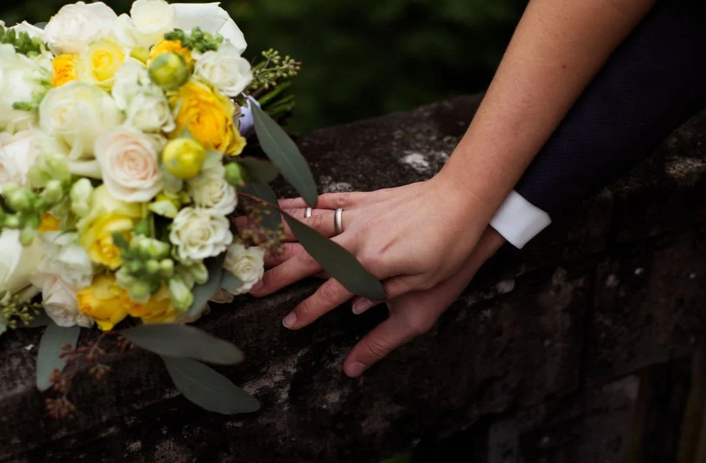 Hände einer Person, die auf einer dunklen Steinmauer liegen, neben einem Brautstrauß aus weißen und gelben Blumen, bei einer Hochzeitsfeier.