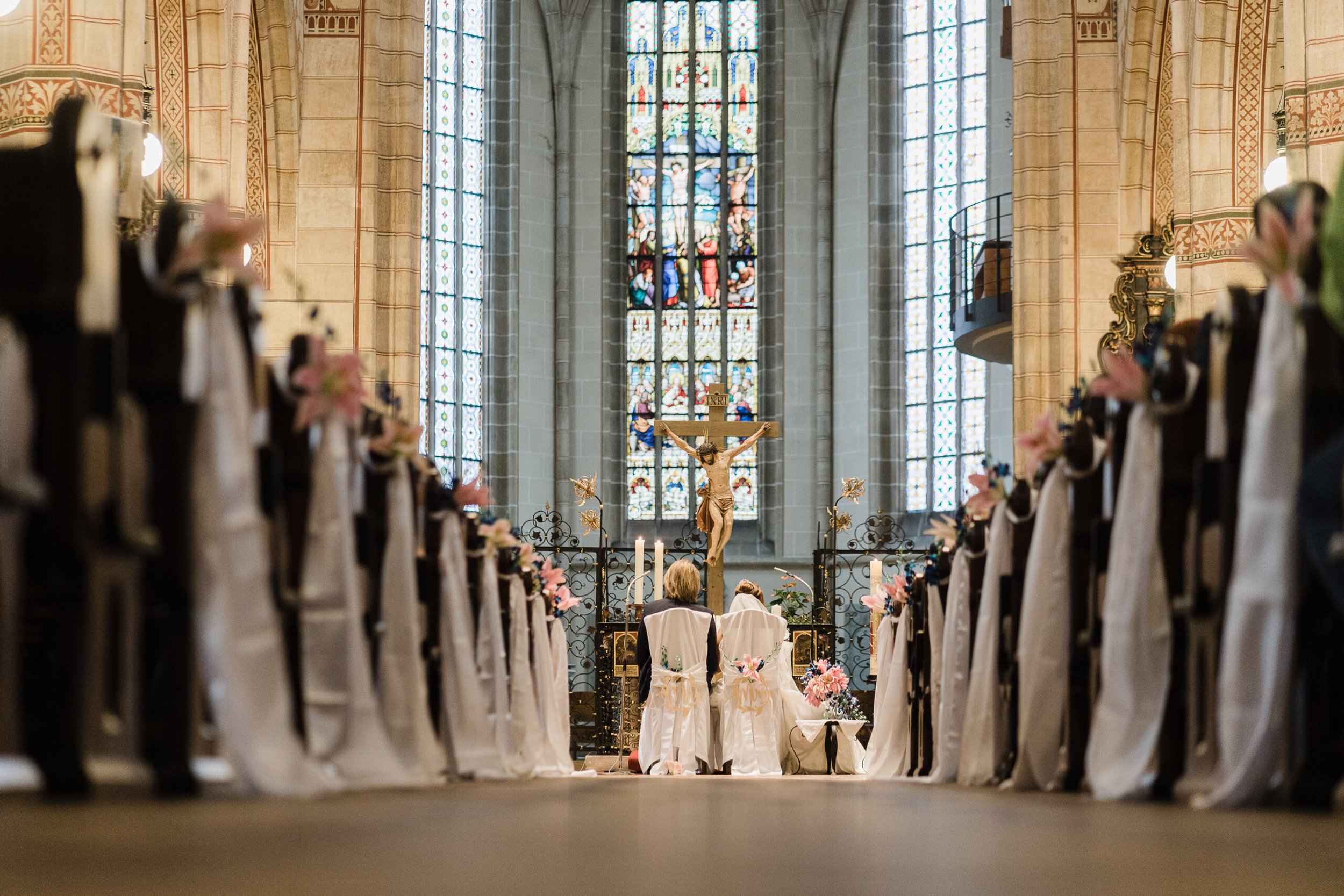 Hochzeitszeremonie in einer Kirche mit Blick auf das Brautpaar vor einem Kreuz und einer bunten Kirchenfenster im Hintergrund.