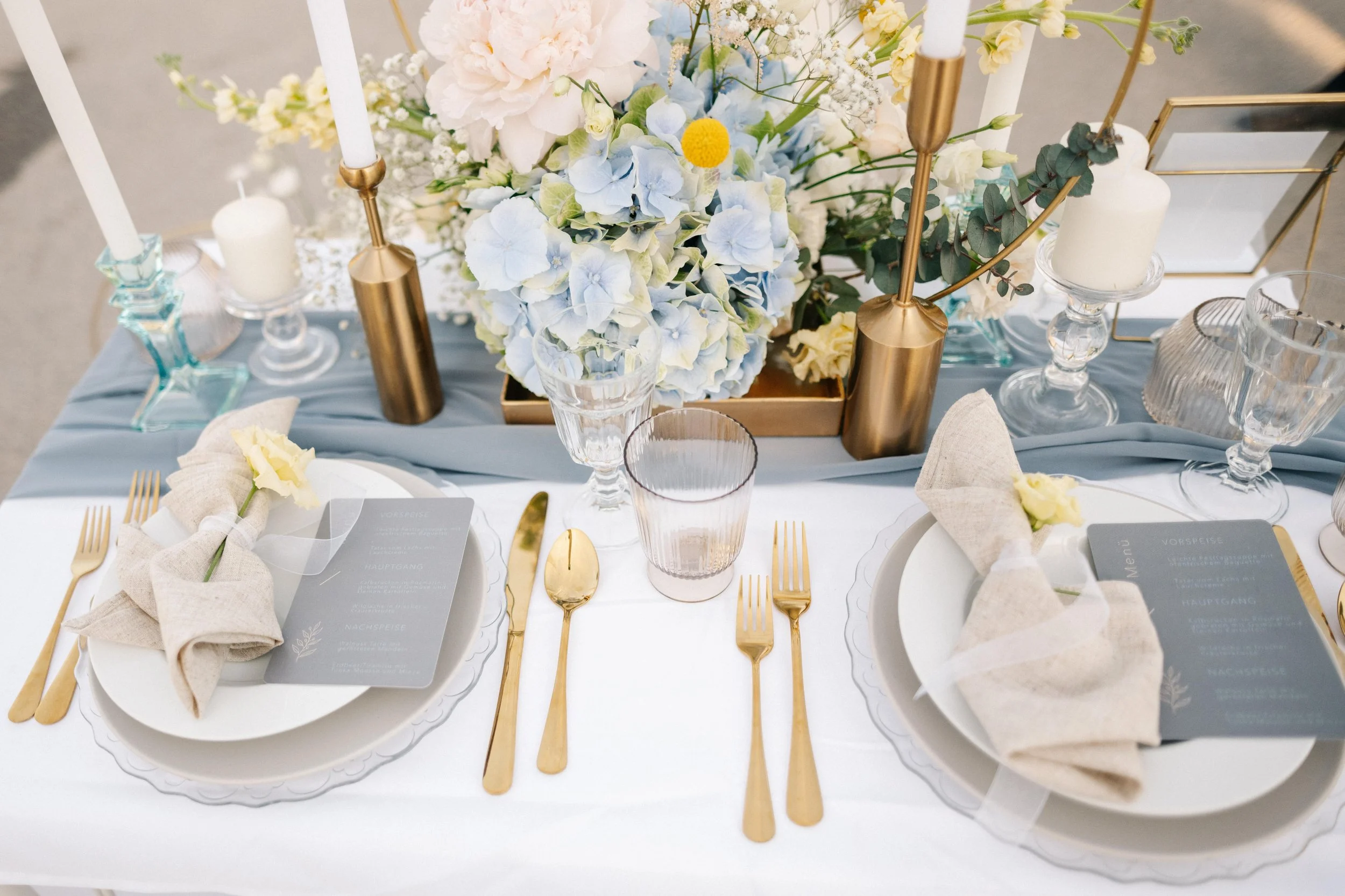 Eleganter Tisch mit weißen Tellern, goldfarbenem Besteck, gefalteten Stoffservietten mit weißen Blumen, und einem eleganten Blumenarrangement in Pastellfarben mit blauen Hortensien und weißem und gelbem Blumenschmuck, umgeben von Kerzen und Dekoratio