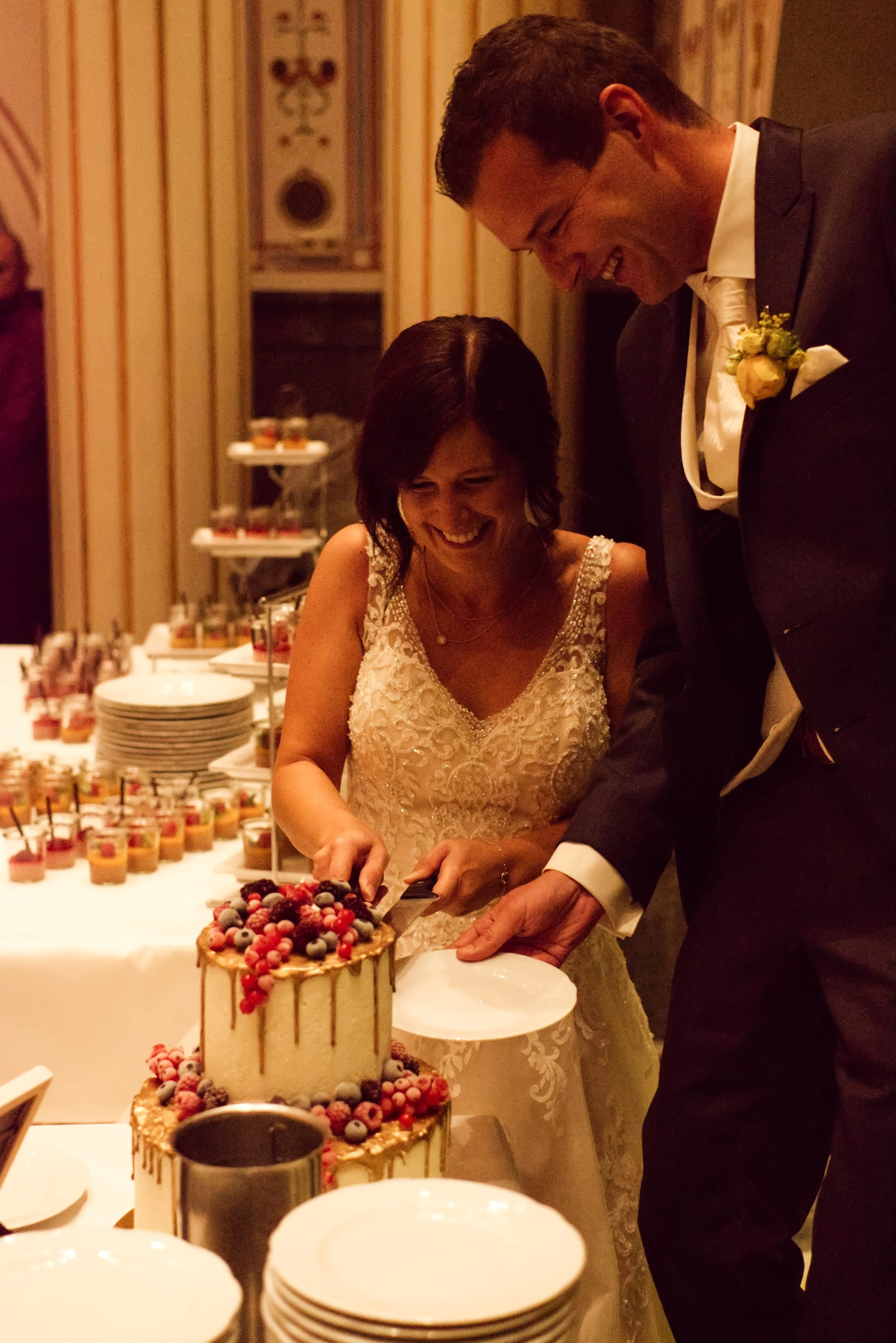 Ein Brautpaar schneidet gemeinsam eine Hochzeitstorte, die mit Beeren und Golddekorationen verziert ist, während sie lachen.