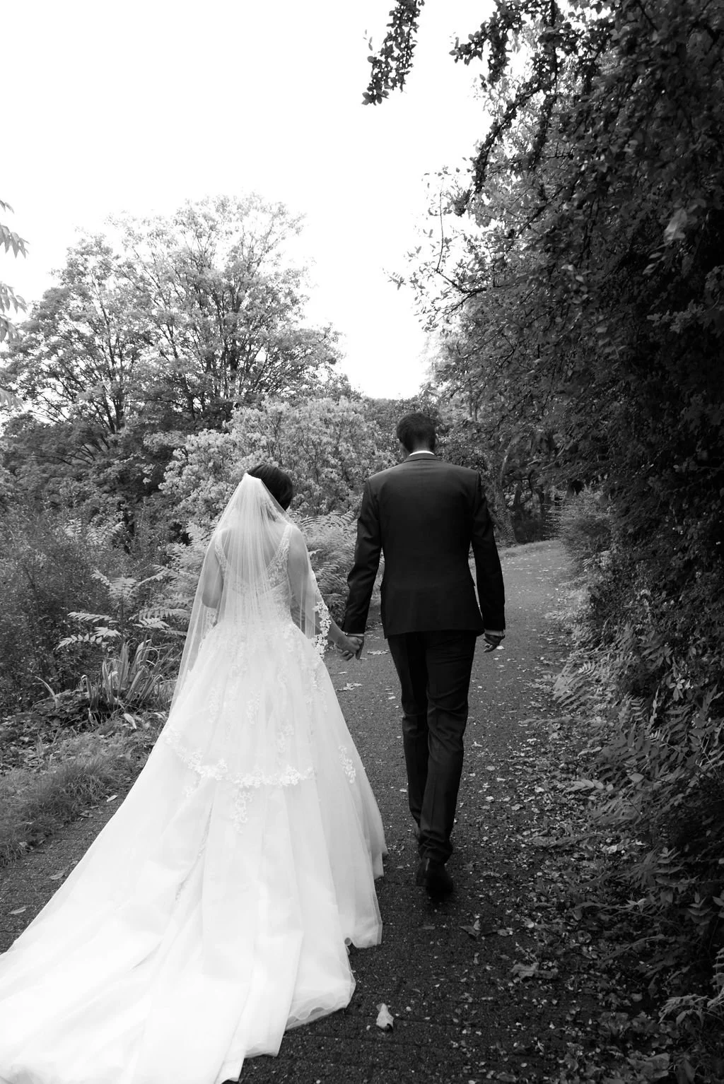 Braut und Bräutigam gehen Hand in Hand auf einem Fußweg in einem parkähnlichen Bereich, umgeben von Bäumen und Sträuchern, in schwarz-weiß