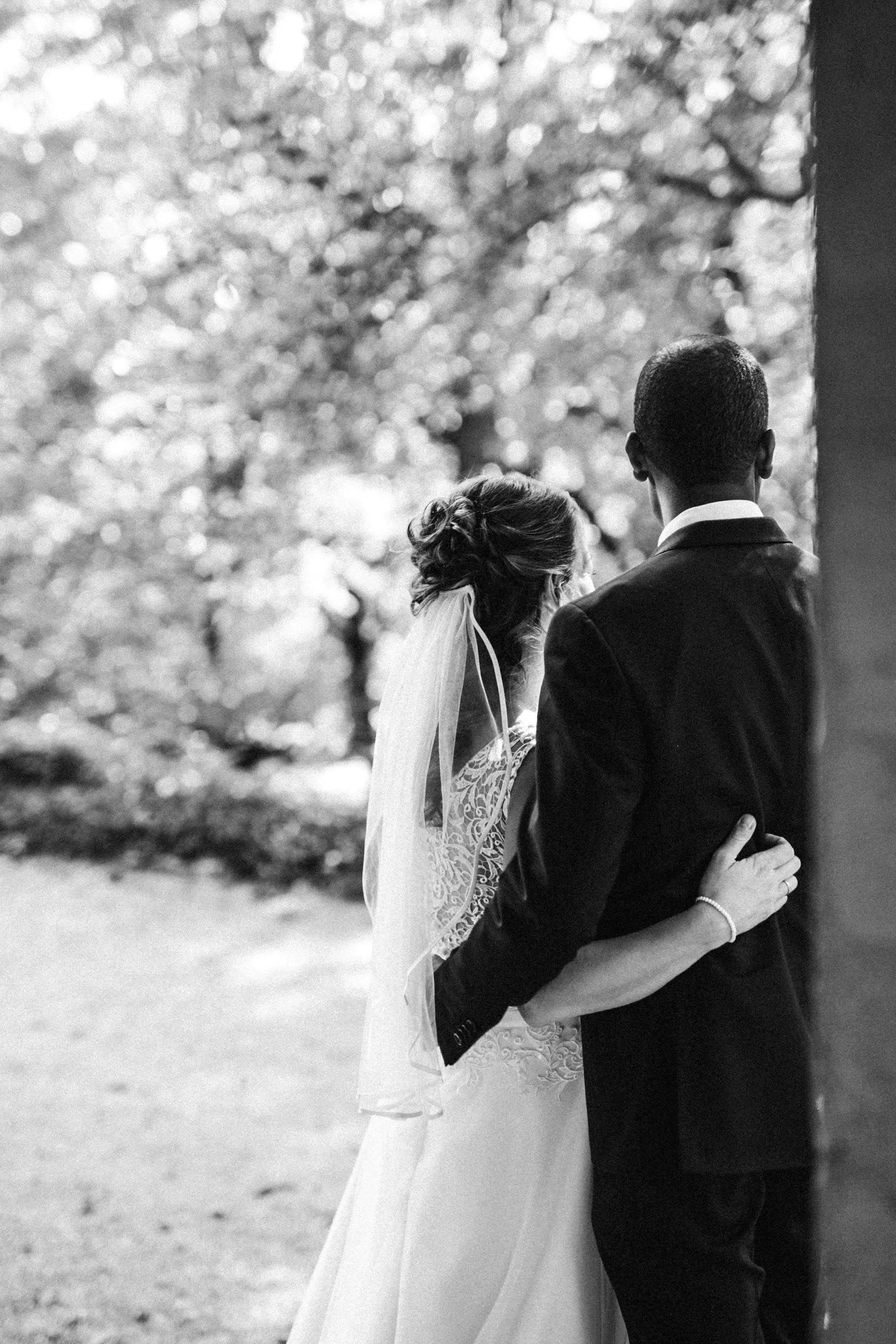 Braut und Bräutigam stehen sich bei einer Hochzeit draußen gegenüber, die Braut trägt ein weißes Kleid mit Spitze und eine Schleife im Haar, der Bräutigam trägt einen dunklen Anzug. Sie umarmen sich vor einer Baumkulisse, schwarz-weiß Fotografie.