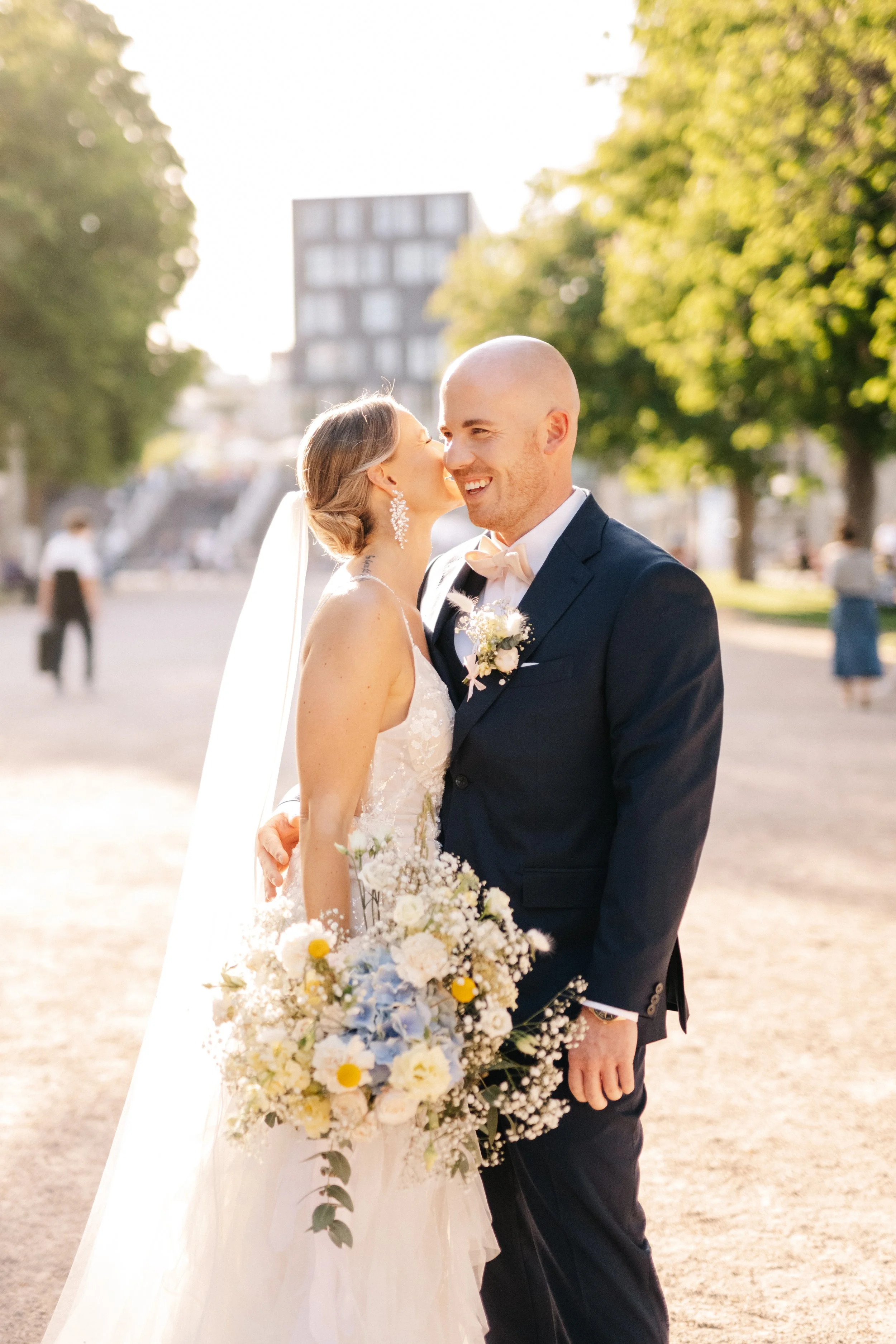 Ein Brautpaar bei einer Hochzeit im Freien, das sich küsst und lächelt, mit einem Blumenstrauß in der Hand, umgeben von grünen Bäumen und warmem Sonnenlicht.