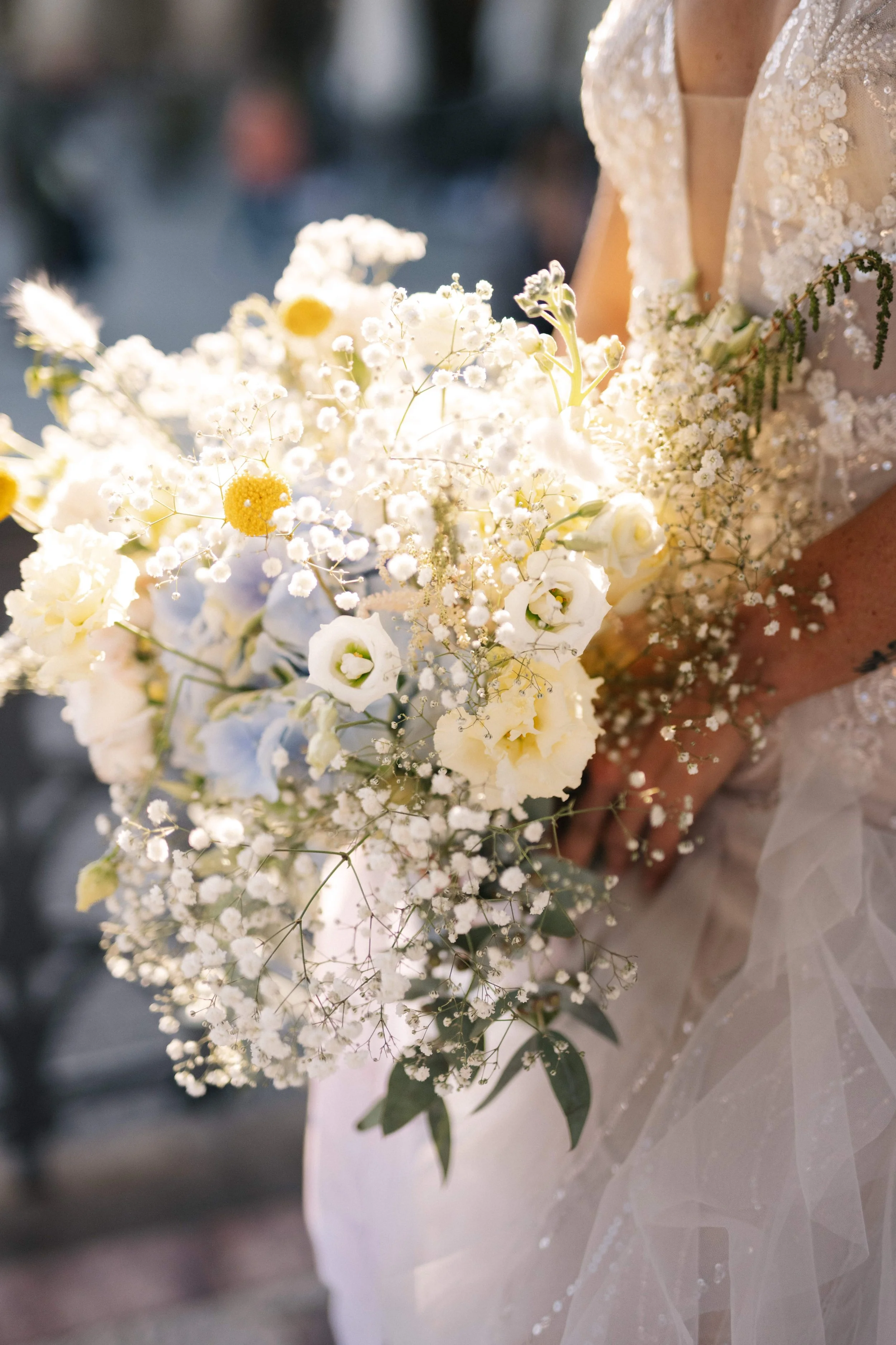 Eine Braut hält einen Blumenstrauß mit weißen und gelben Blumen, umgeben von zarten kleinen Blüten, bei einer Hochzeit im Freien.