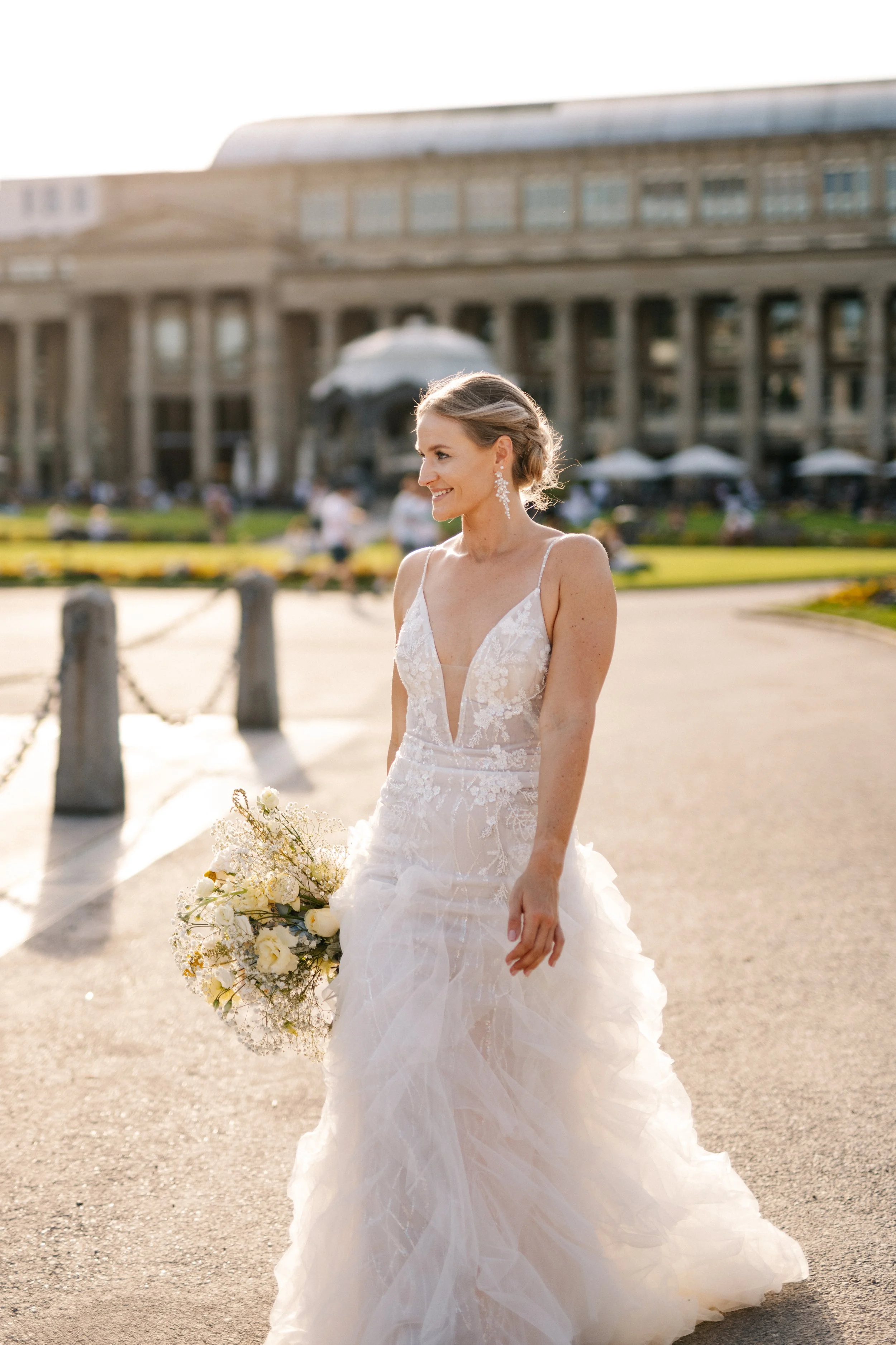 Braut in hochzeitskleid mit Blumenstrauß vor einem historischen Gebäudekomplex bei Sonnenuntergang.
