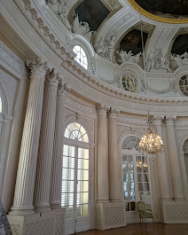 Innenraum eines historischen, prunkvollen Raums mit großen Fenstern, weißen Säulen und einer goldfarbenen Kronleuchter