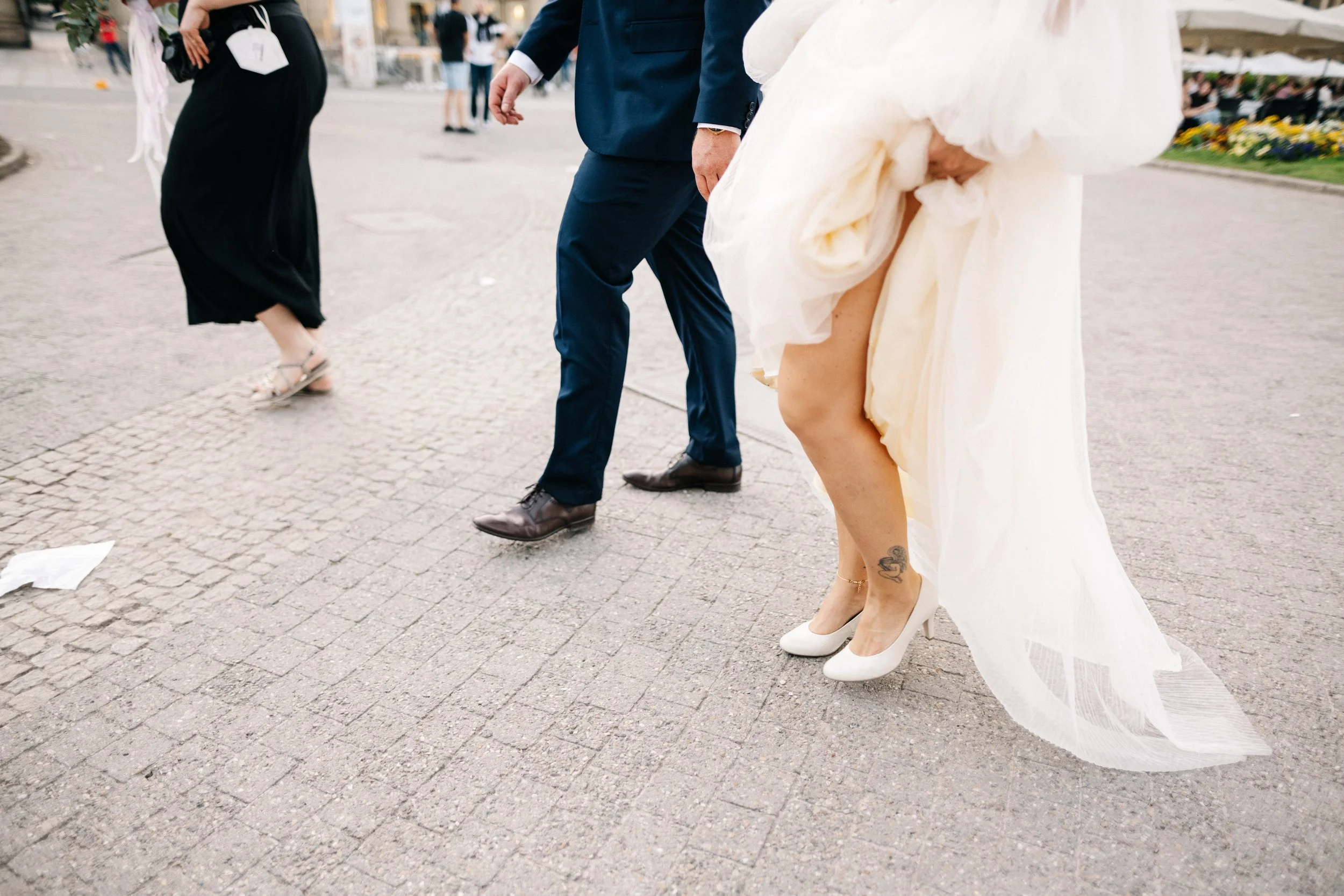 Menschen in formeller Kleidung, darunter eine Braut in einem weißen Kleid mit hohen Schuhen und einem Tattoo am Bein, laufen auf einer gepflasterten Fläche bei einer Veranstaltung im Freien.
