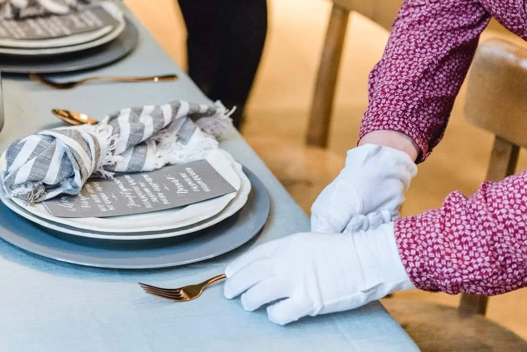 Eine Person richtet einen gedeckten Esstisch mit weißen Handschuhen. Es gibt Teller, Besteck und eine dankende Karte auf einem gestreiften Tuch.
