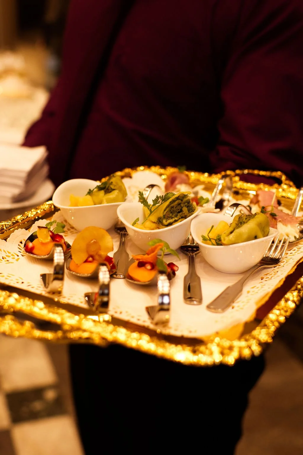 Ein Service mit kleinen Vorspeisen und Häppchen auf einem goldfarbenen Tablett, gehalten von einer Person in dunklem rotem Jackett.