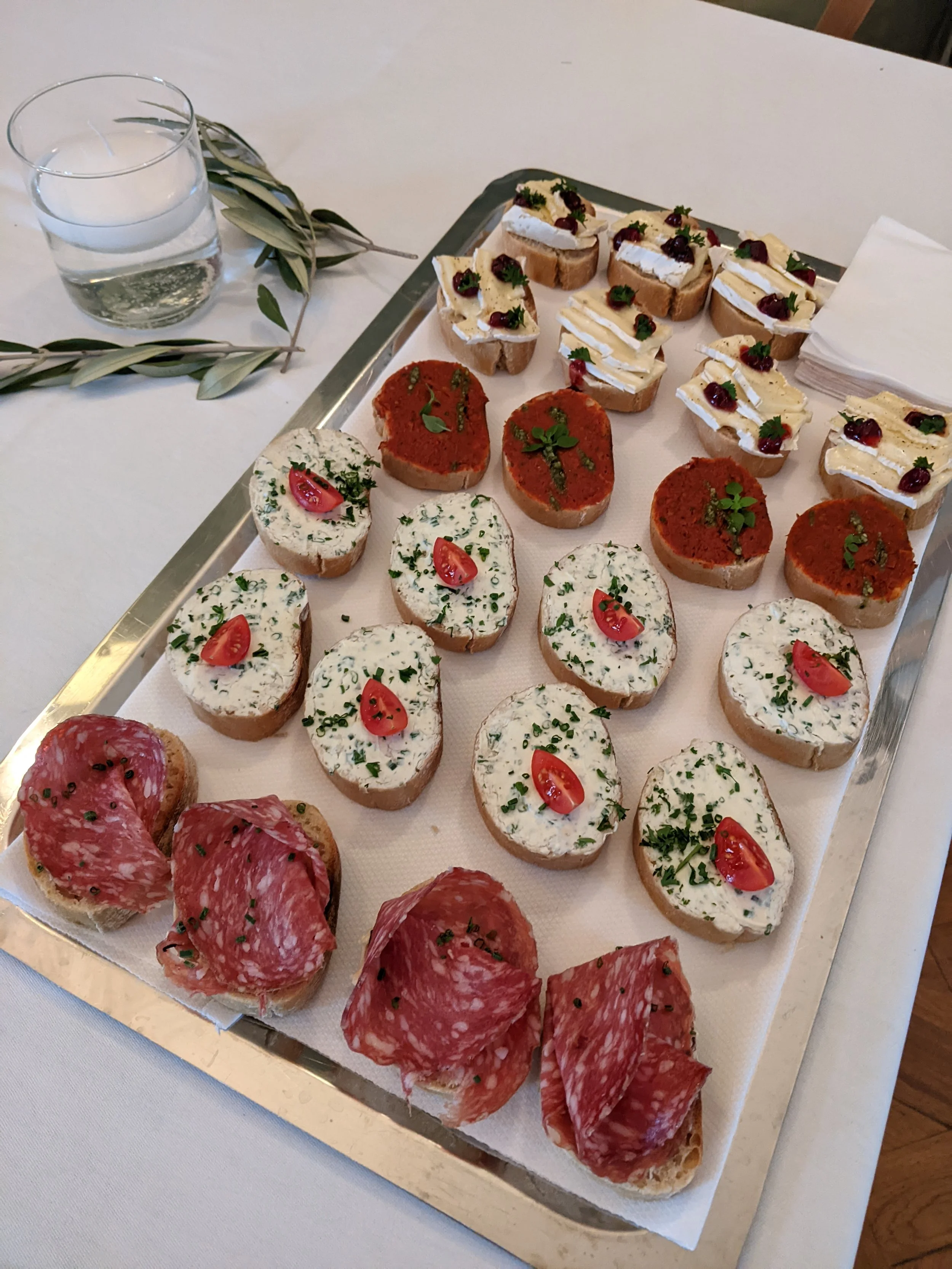 Verschiedene belegte Brote auf einem Serviertablett, inklusive Tomaten, Kräuter, Wurst und Käse, auf einer weißen Tischdecke.