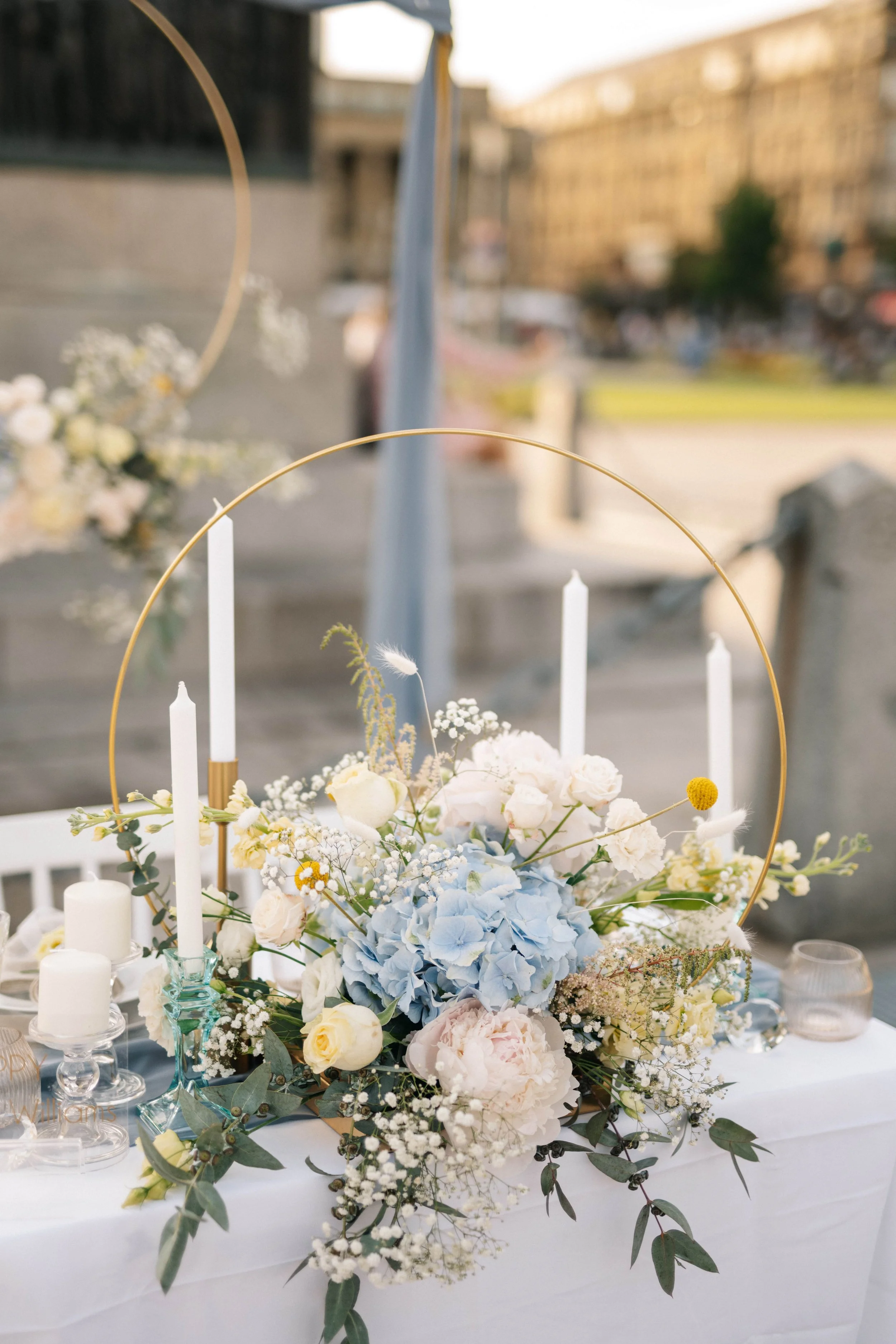 Blumenarrangement mit weißen und blauen Blumen, weiße Kerzen und Dekoration auf einem Tisch bei einer Veranstaltung im Freien.