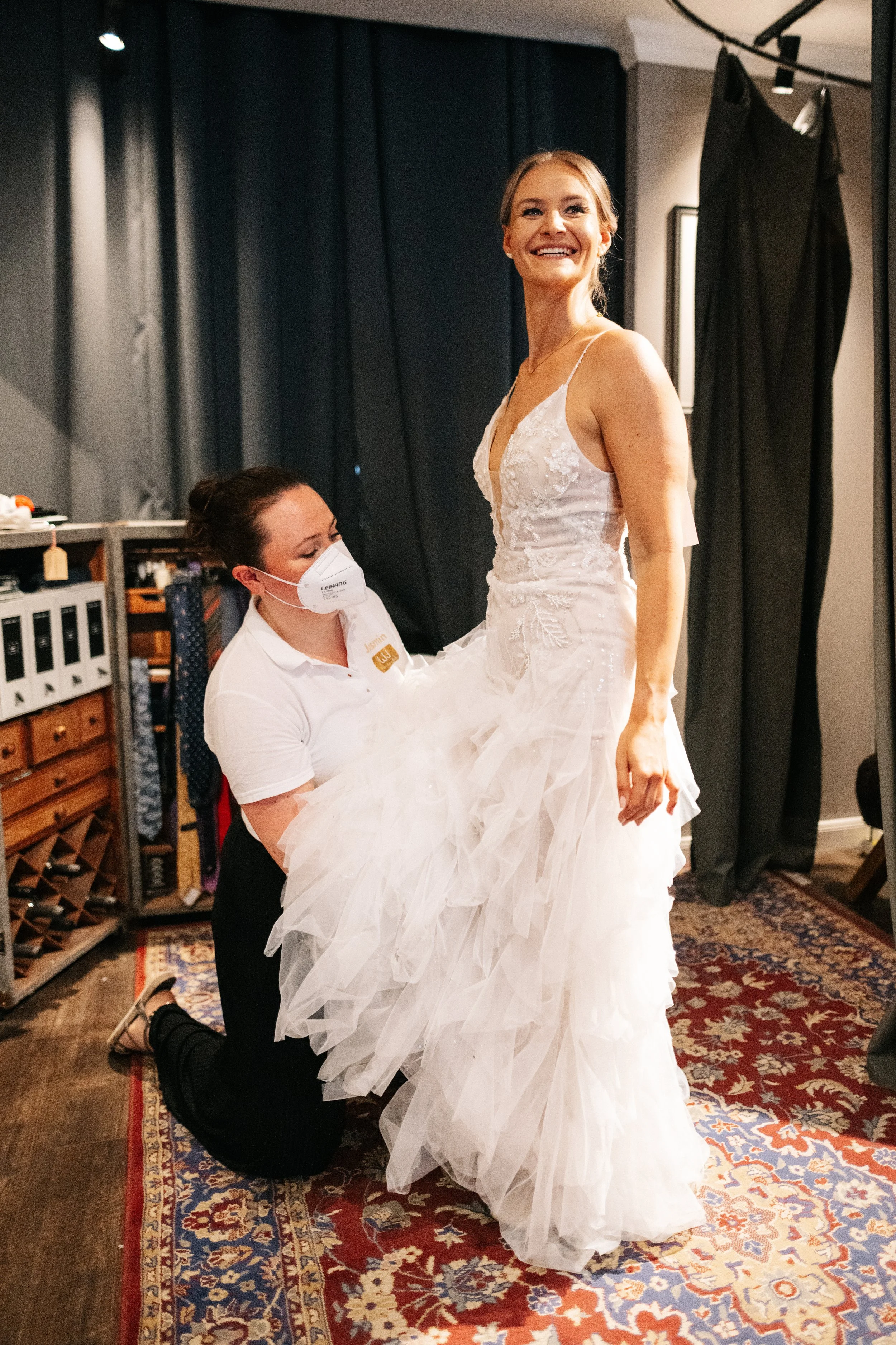 Frau probiert ein Brautkleid in einem Schneideratelier, während eine Mitarbeiterin sie an einem Knien anfasst.