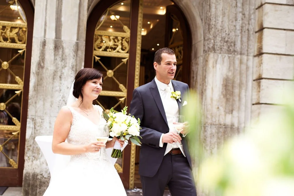 Ein Brautpaar bei einer Hochzeitszeremonie draußen vor einem historischen Gebäude mit großen Türen und goldverzierten Fenstern. Die Braut trägt ein weißes Brautkleid, hält einen Blumenstrauß und ein Glas Wein. Der Bräutigam trägt einen dunklen Anzug 