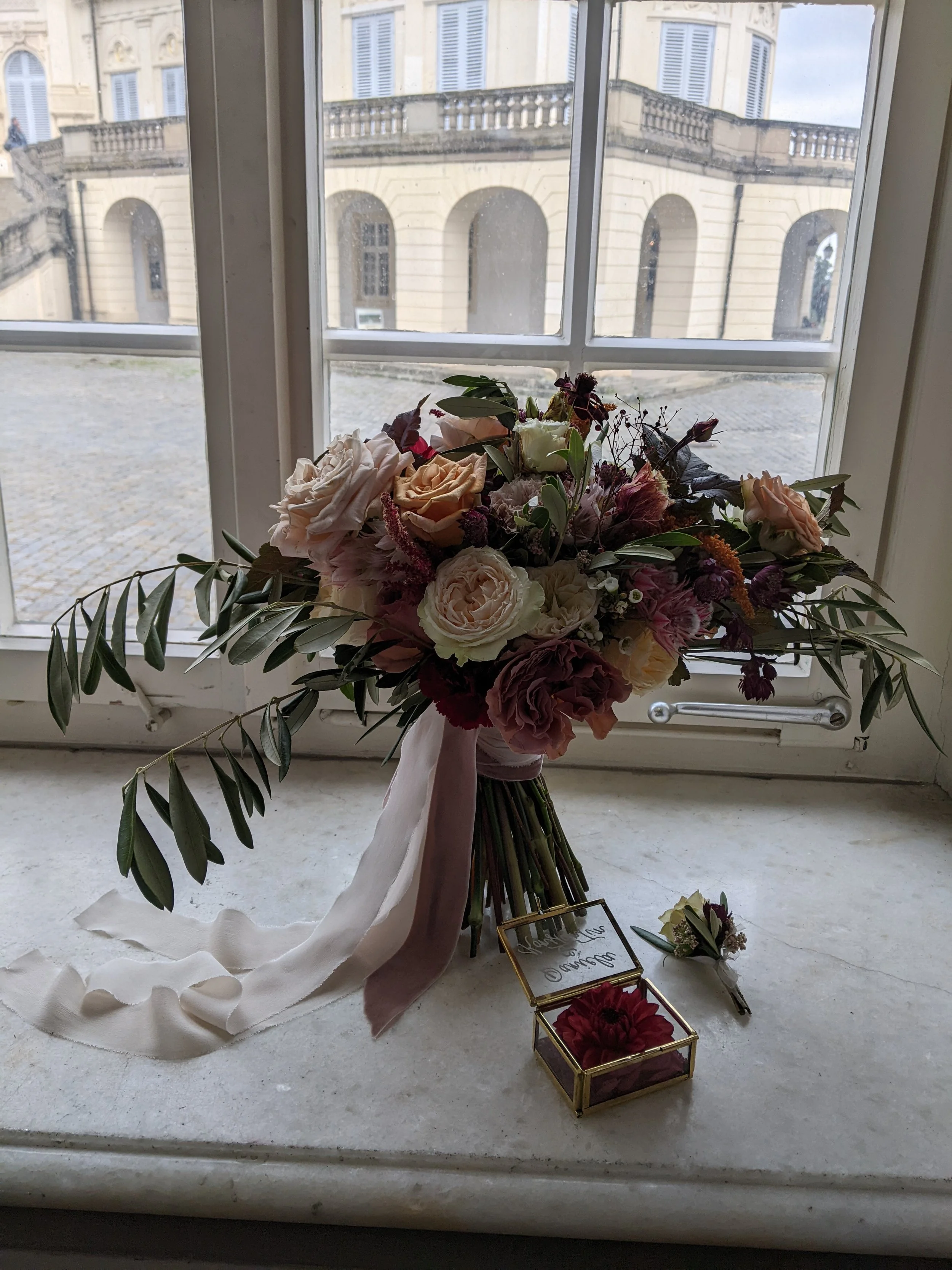 Ein Blumenstrauß mit Rosen und anderen Blumen auf einem Fensterbrett, daneben eine kleine Box mit roten Blumen und eine kleine Blume, im Hintergrund ein Blick auf ein altes Gebäude mit Rundbögen.