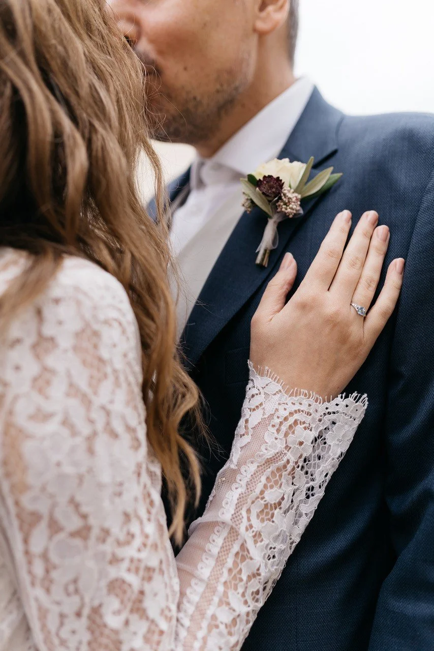 Ein Brautpaar bei einer Hochzeitsbegegnung, Nahaufnahme von Hände und Oberkörper, die Braut trägt ein Spitzenkleid, zeigt einen Verlobungsring, der Bräutigam trägt Anzug mit Ansteckblume.