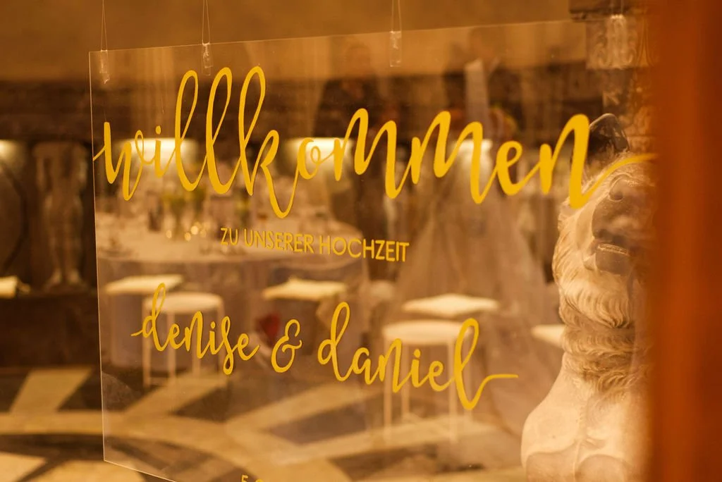 Willkommensschild für eine Hochzeit mit Namen Denise und Daniel, in einem festlichen Raum mit antiken Möbeln und Skulpturen.