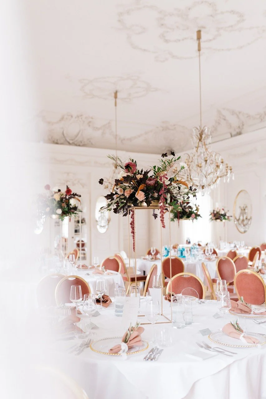 Eleganter festlicher Saal mit runden Tischen, blumigen Mittelstücken, Stühlen mit rosa Polstern, Kristall-Kronleuchtern und elegante Dekorationen.