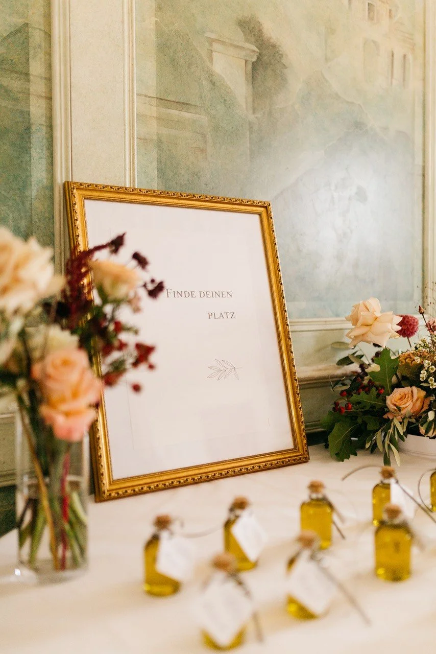 Ein goldgerahmtes Schild mit dem deutschen Text 'Finde deinen Platz' ist auf einem Tisch mit Blumen und kleinen Flaschen mit Öl oder Parfüm platziert.