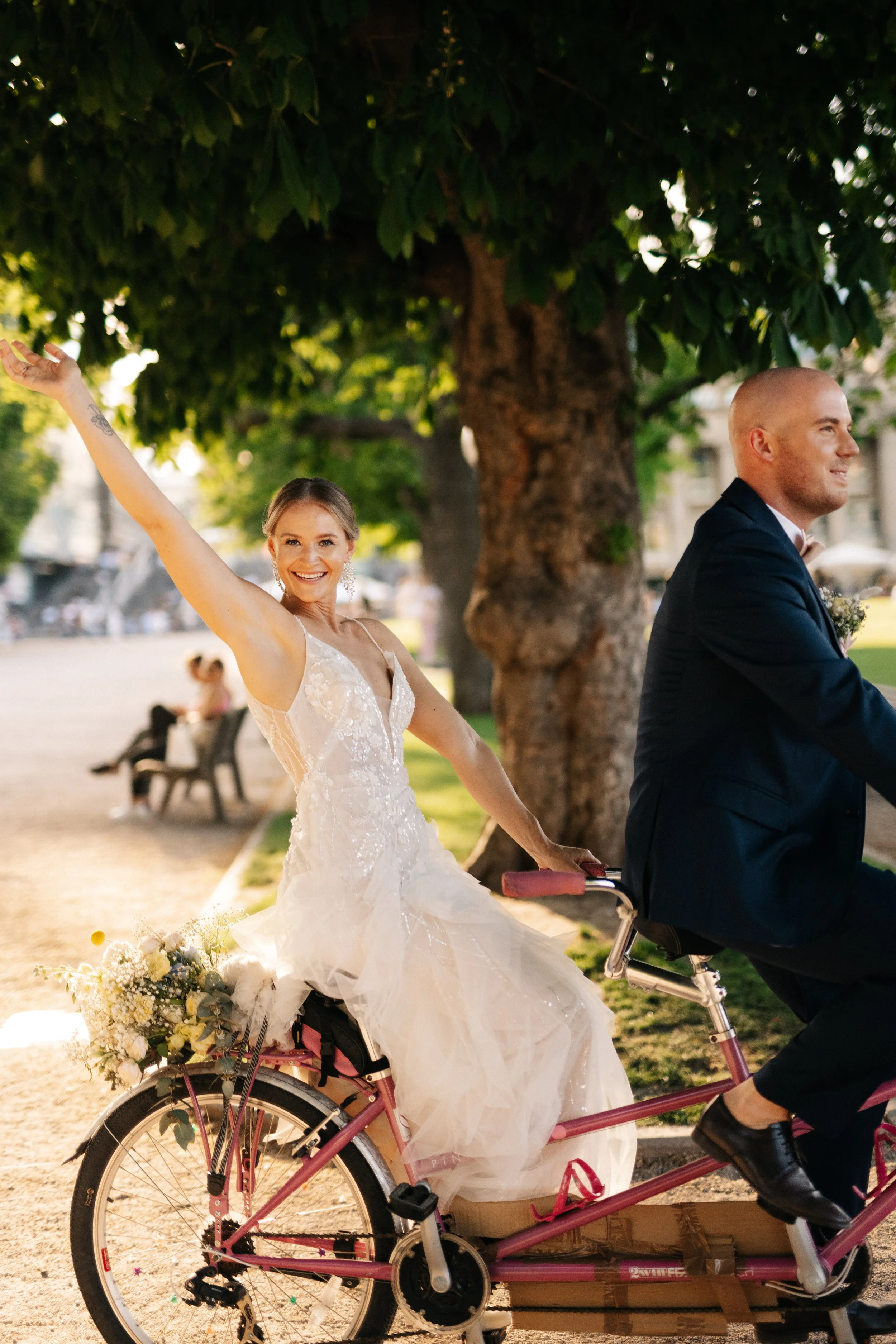 Braut in weißem Kleid sitzt auf Fahrrad, das von Bräutigam gefahren wird, im Park bei Sonnenuntergang, mit glücklichem Gesicht und Blumen im Korb