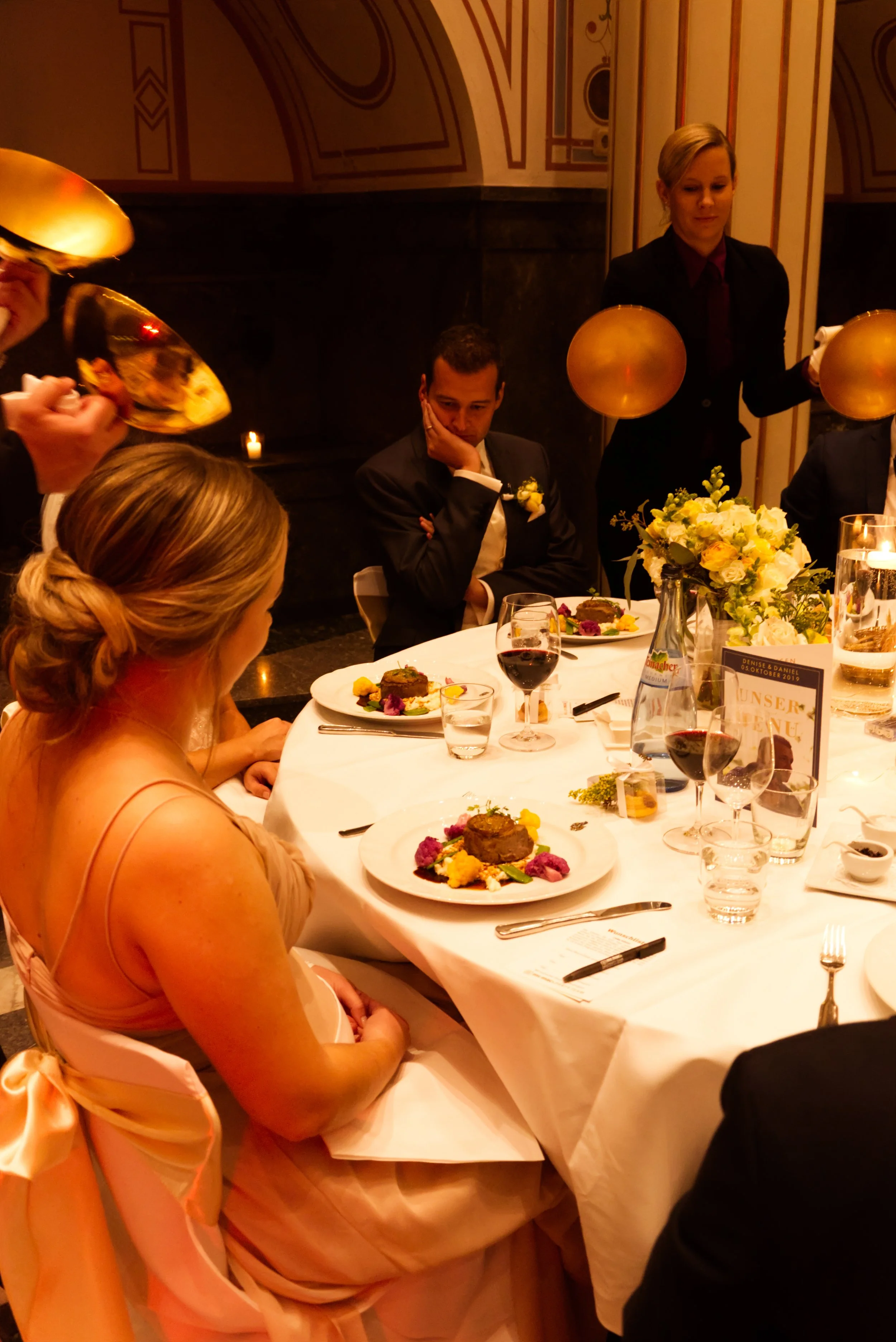 Menschen bei einem festlichen Abendessen, einige mit goldenen Tellern, in einem dekorierten Raum, serviert von Kellnerinnen.