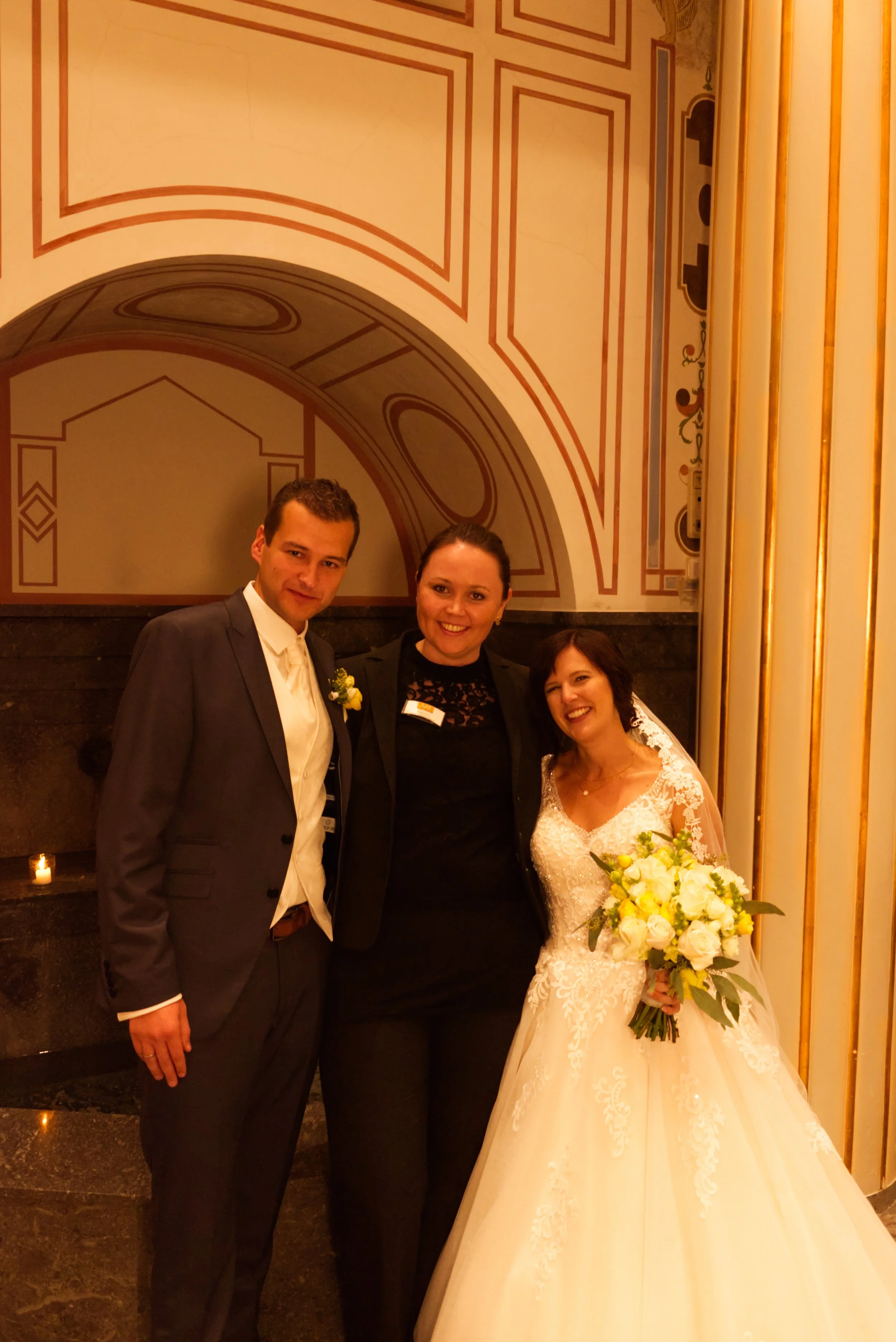 Ein Brautpaar mit einer Begleitperson bei einer Hochzeit, alles lächelnd, im Innenraum mit goldenen Verzierungen und einer hohen Decke, Braut trägt ein weißes Hochzeitskleid und hält einen Blumenstrauß.