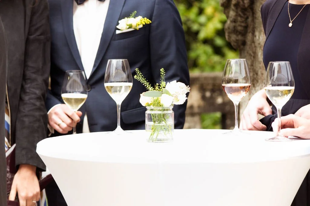 Menschen bei einer Feier mit Weingläsern auf einem Stehtisch, im Hintergrund grüne Pflanzen, eine kleine Blumenvase auf dem Tisch.