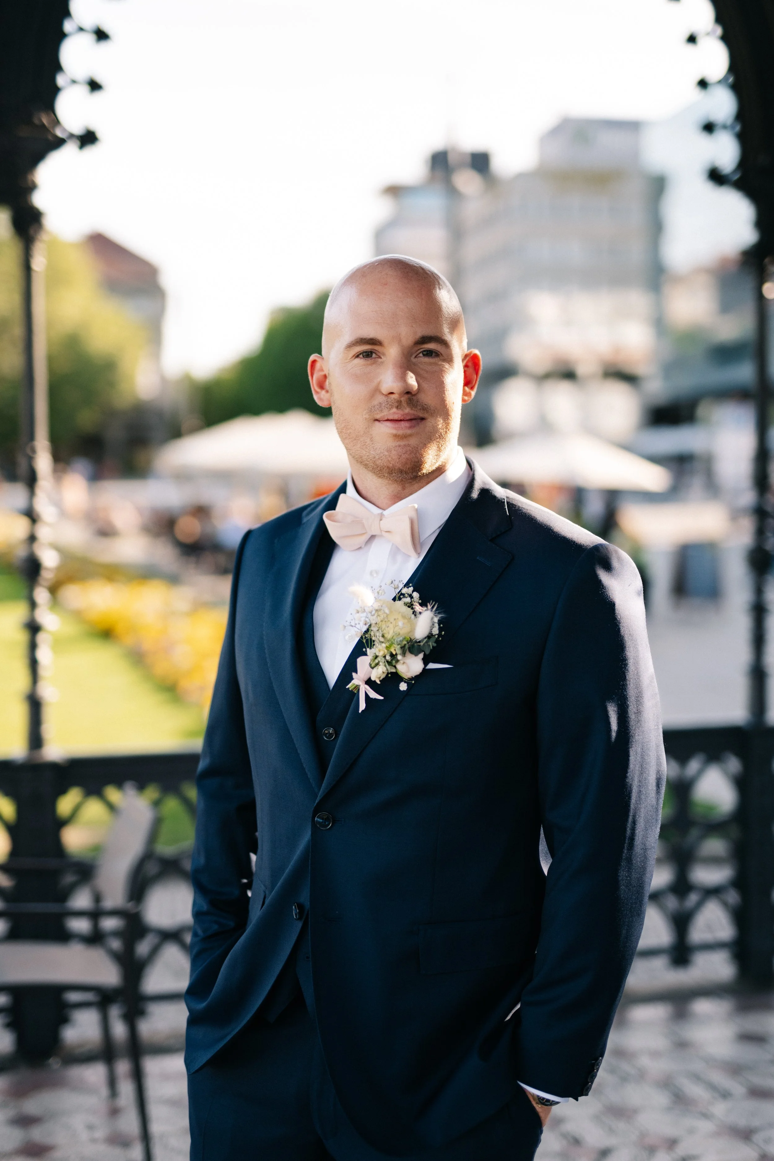 Junger Mann in formeller schwarzer Anzug mit weißem Hemd und Blumenanstecker, stehend im Freien mit Stadt- und Parkhintergrund, wahrscheinlich an einem besonderen Tag wie Hochzeit.