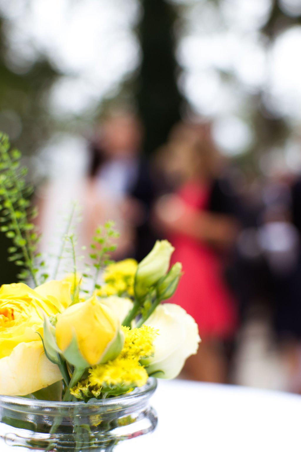 Nahaufnahme eines Blumenstraußes mit gelben und weißen Blumen in einem Glasvasen, im Hintergrund unscharfe Menschen bei einer Veranstaltung.
